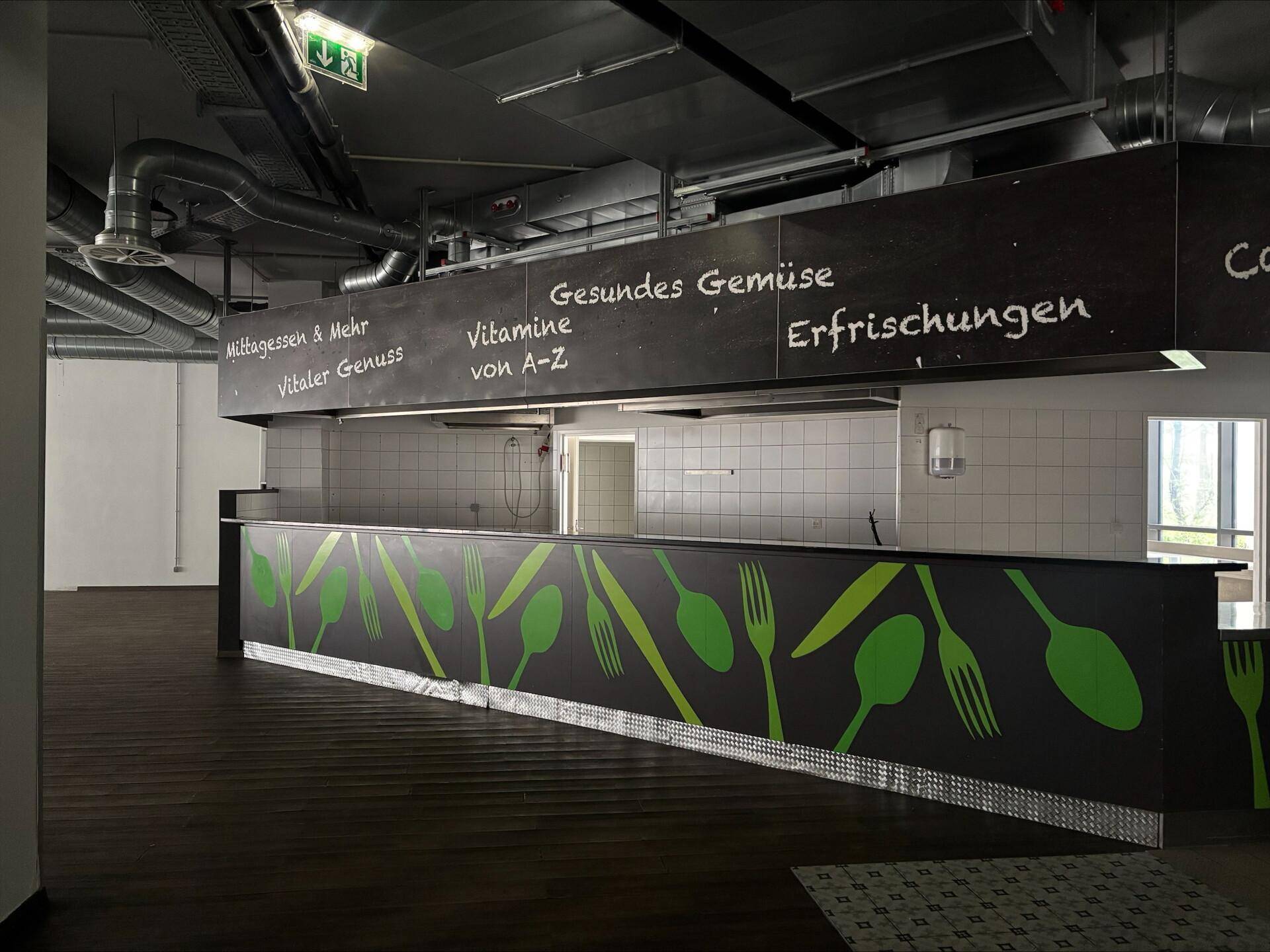 Kantinenbereich mit Theke, Beschriftungen und sichtbaren Lüftungsanlagen an der Decke.