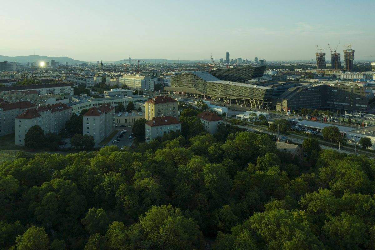 Panoramablick über eine Stadt mit ausgedehnten Grünflächen, Gebäuden und Bergen im Hintergrund.