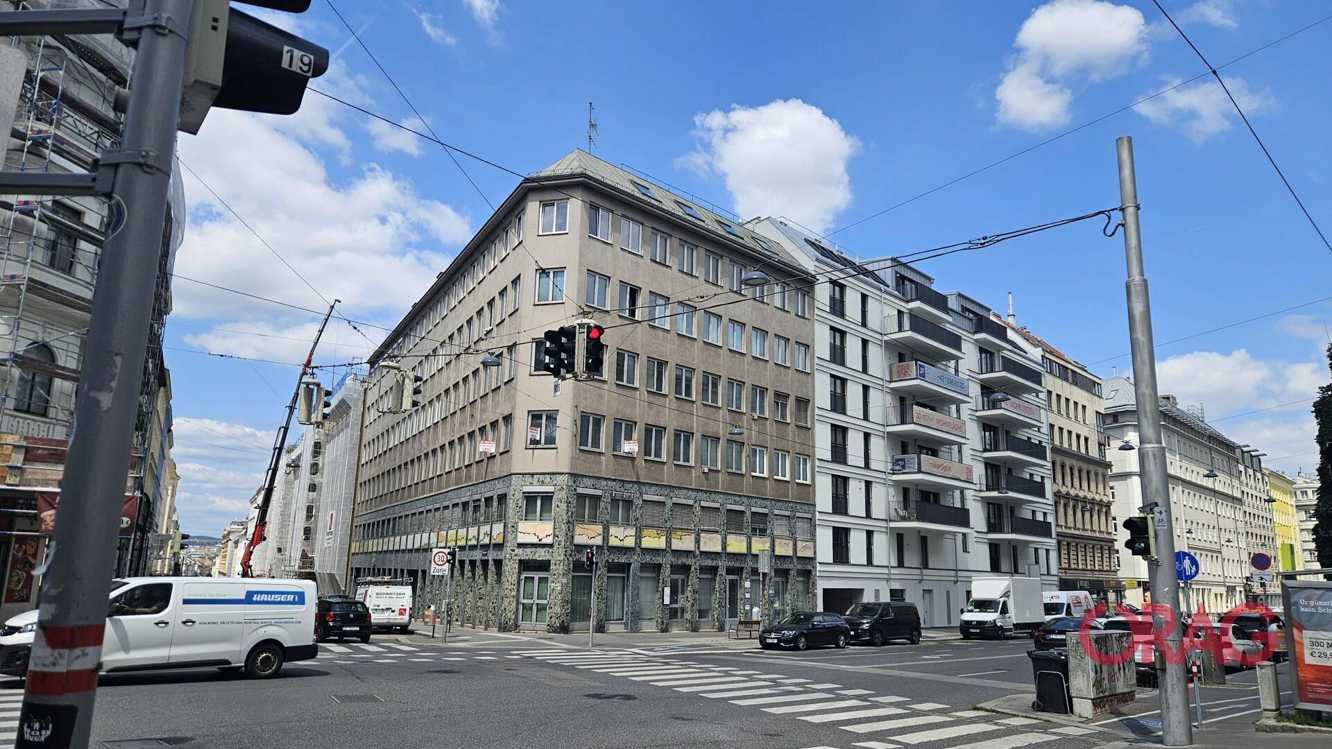 Eckgebäude in einer belebten Stadtlandschaft mit angrenzenden modernen Gebäuden und Straßenverkehr bei Tageslicht.