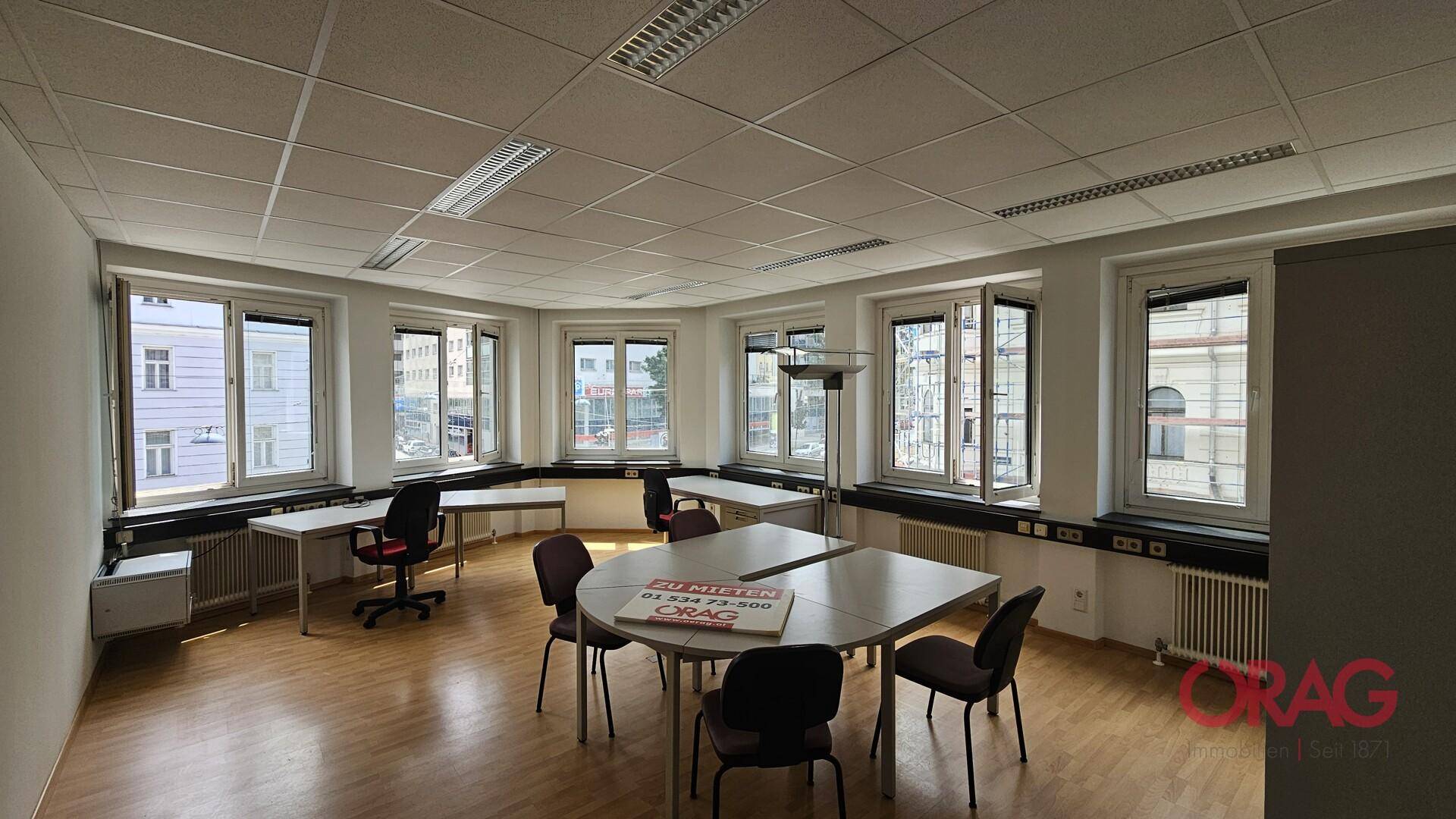 Geräumiges Eckbüro mit zahlreichen Fenstern, Holzfußboden und einem ovalen Besprechungstisch.