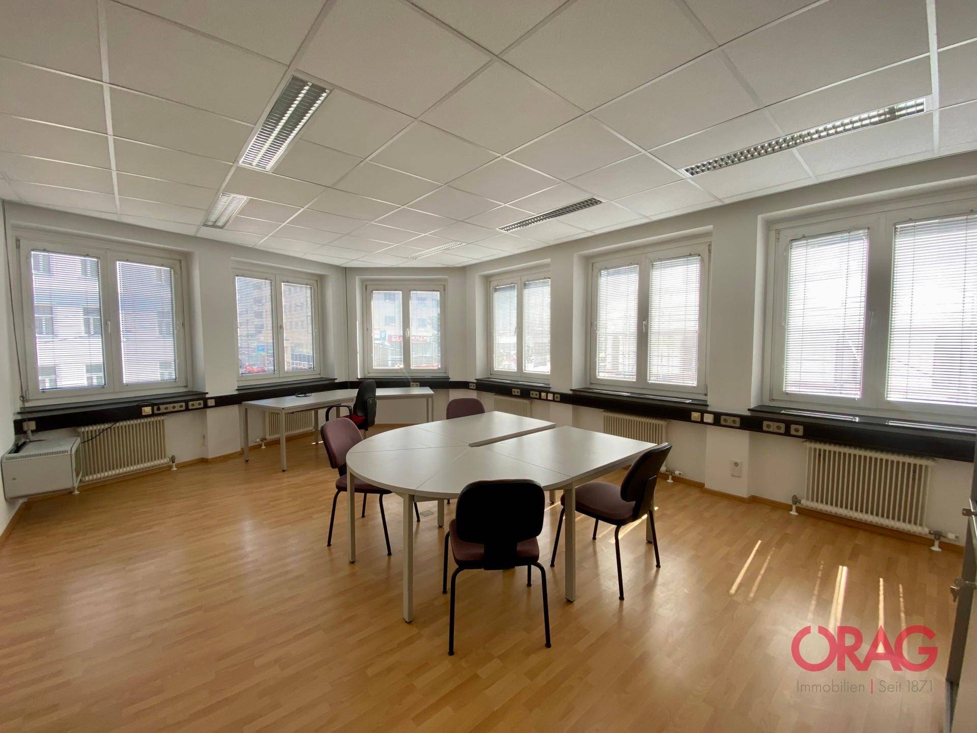 Großes Eckbüro mit vielen Fenstern, Holzfußboden und einem ovalen Konferenztisch.