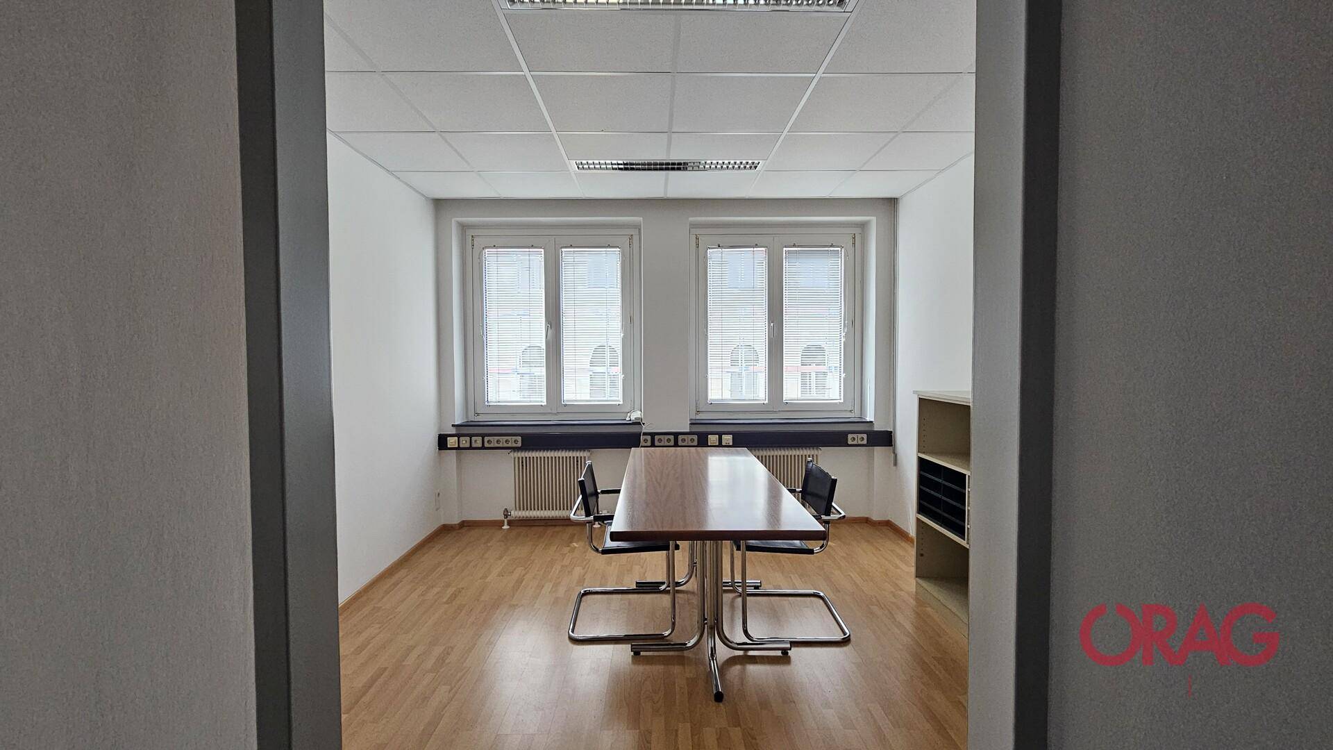 Büro mit Holzboden, zwei Fenstern und einem Konferenztisch, gesehen durch einen Türrahmen.