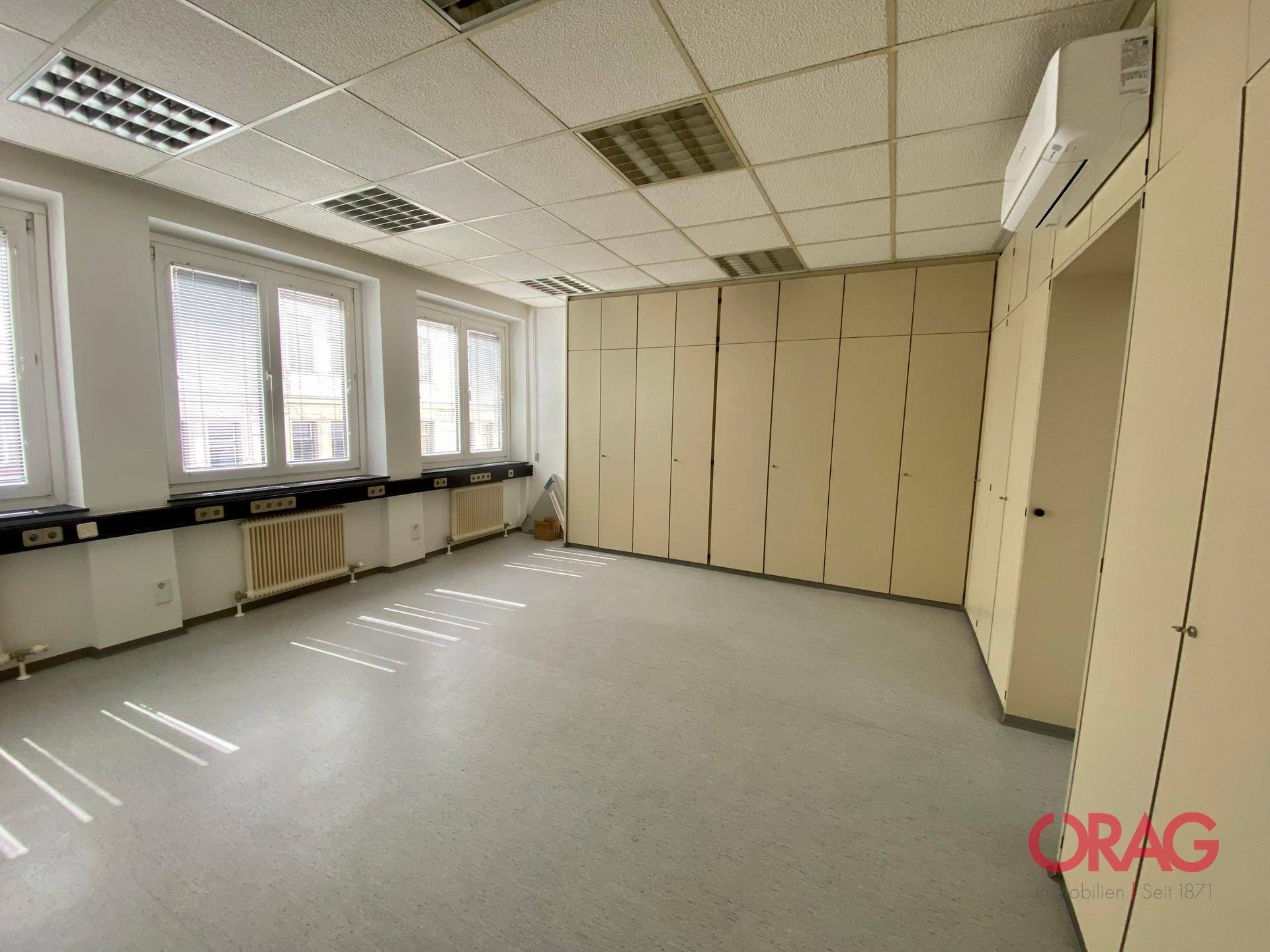 Geräumiger Büroraum mit Vinylboden, Einbauschränken entlang einer Wand und Fenstern.