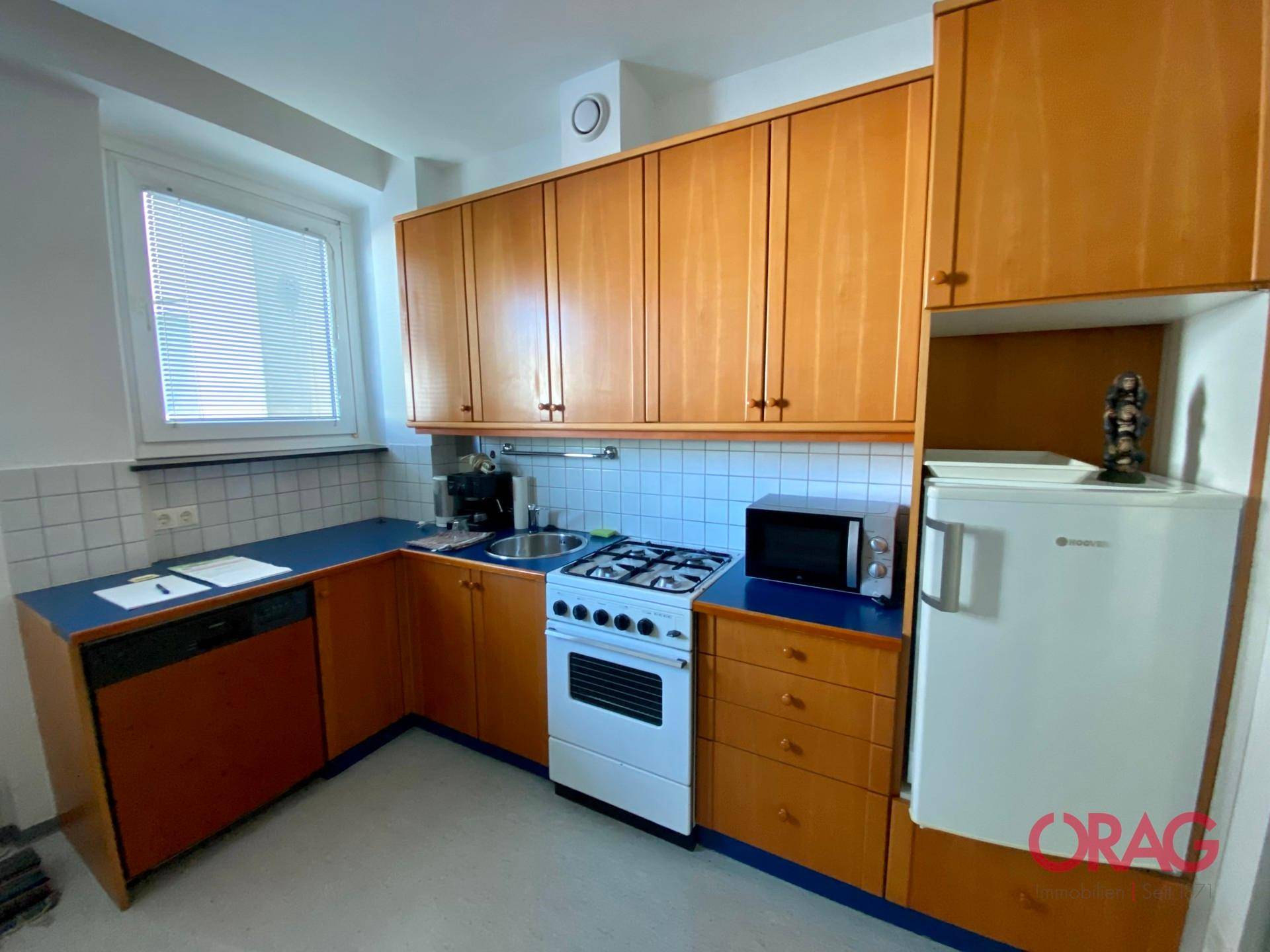 Funktionale Küche mit Holzschränken, Gasherd, Mikrowelle, Kühlschrank und Fenster.