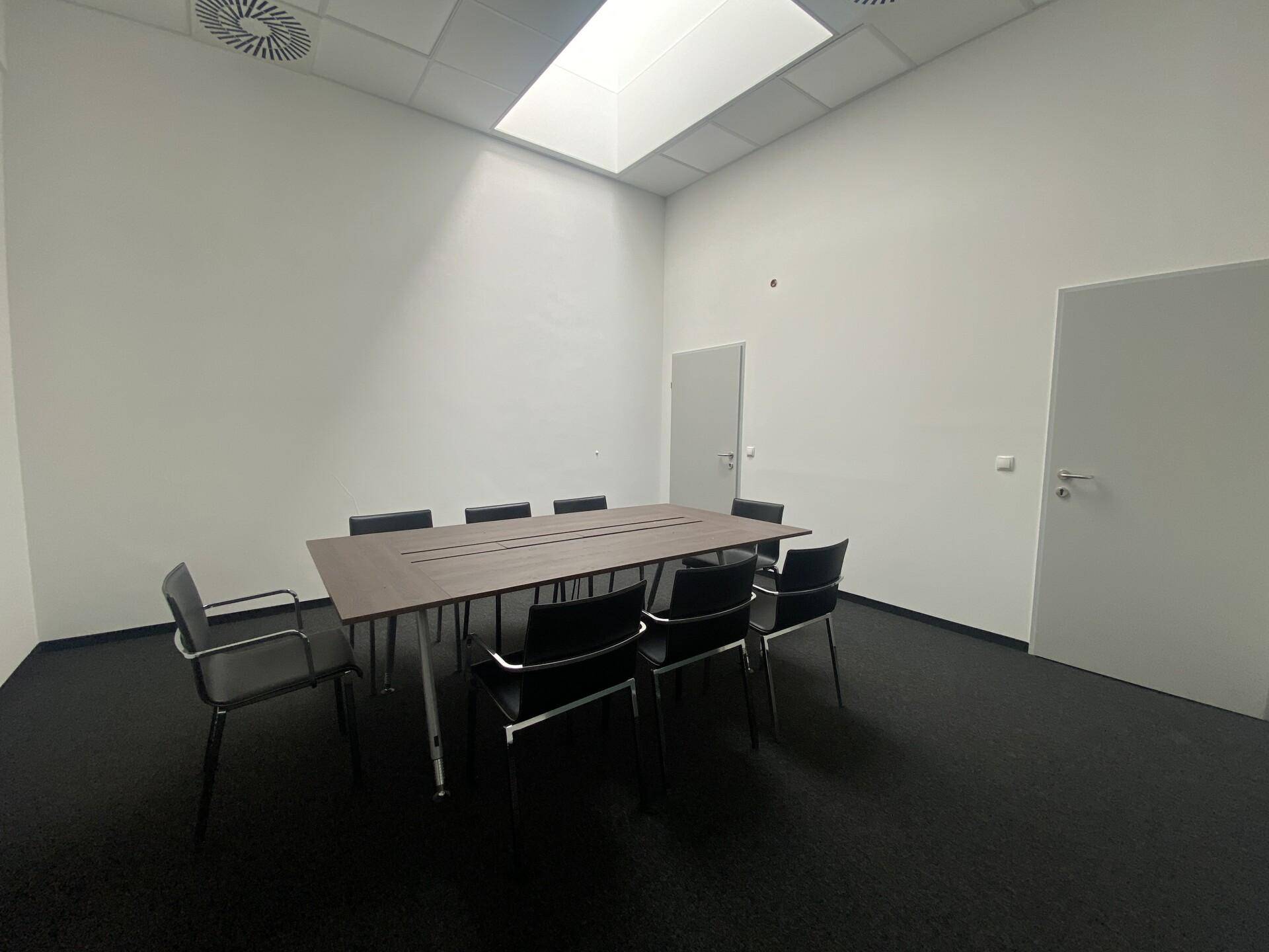 Konferenzraum mit großem Holztisch, schwarzen Stühlen und Teppichboden.