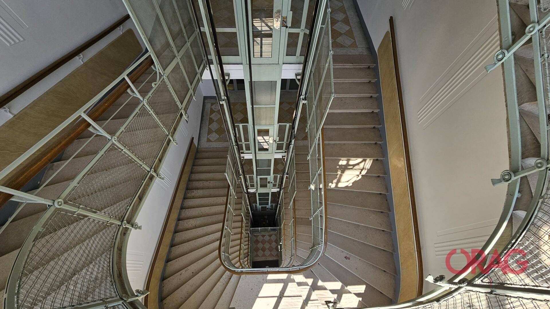 Historisches Treppenhaus mit einem zentralen Aufzugschacht und kunstvollen Metallgeländern, durchflutet von Tageslicht.