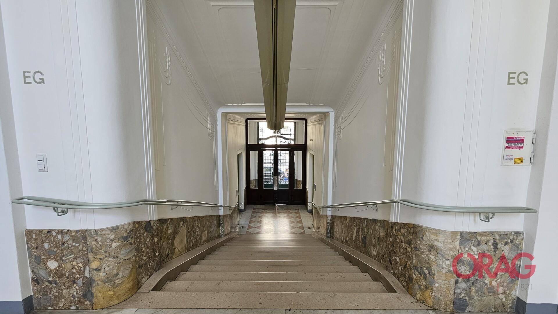 Eingangsbereich mit breiter Steintreppe, Marmorverkleidung an den Wänden und hohen Decken, führt zu einer Doppeltür.