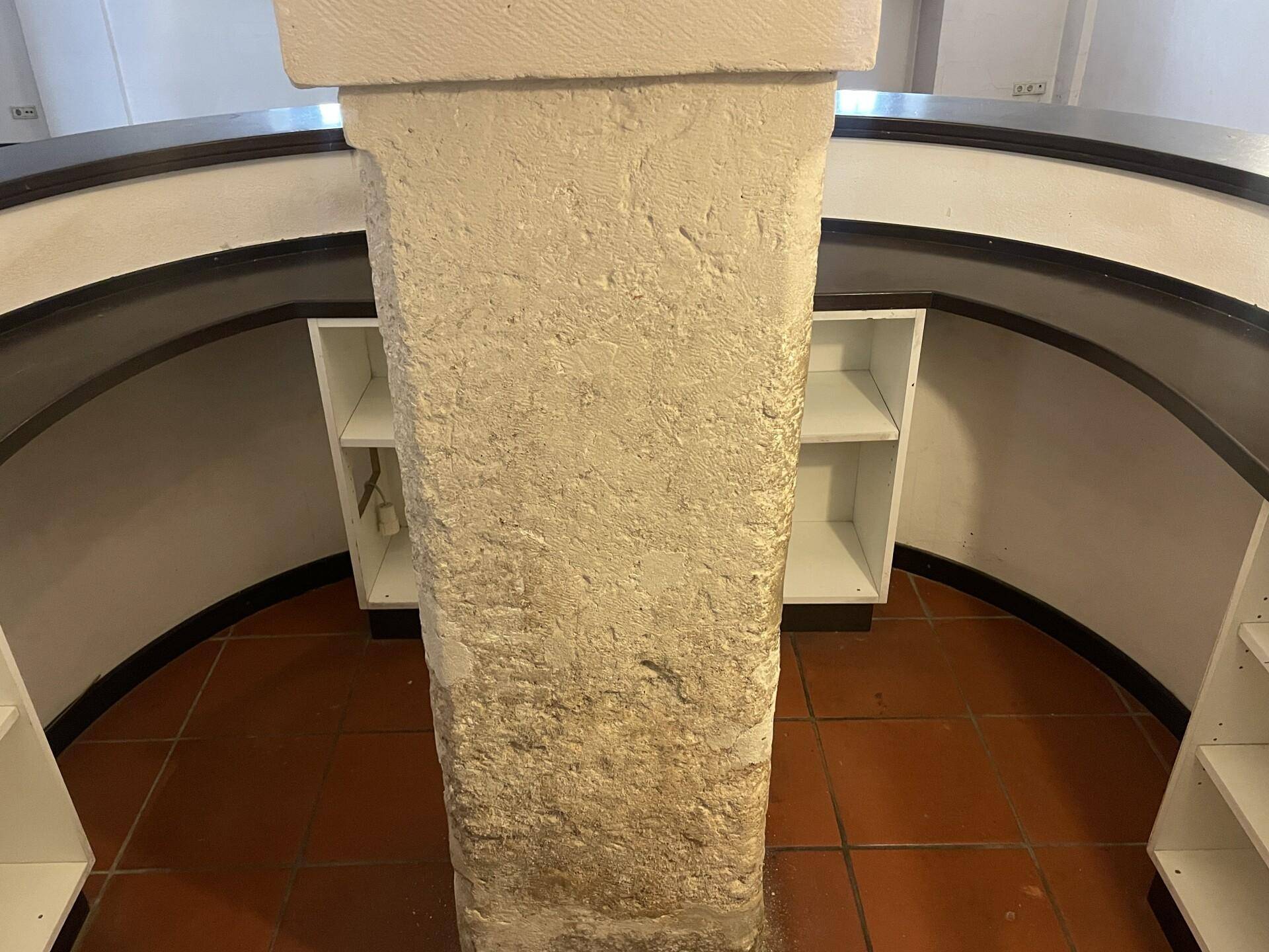 Detailansicht einer Steinsäule in einem Gewölberaum mit Terrakottafliesen und Regalen.