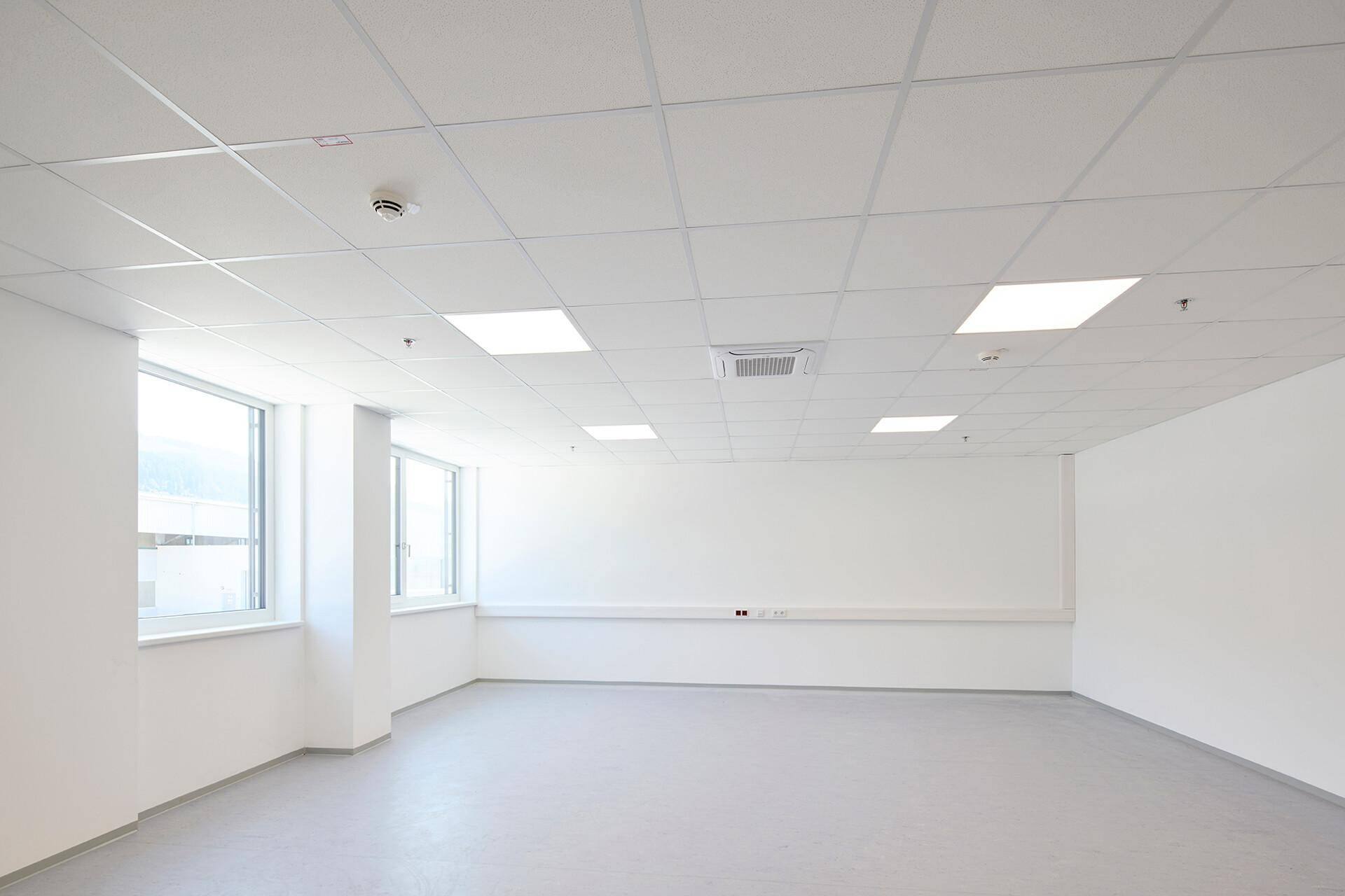 Helles, leeres Büro mit weißen Wänden, Rasterdecke und großen Fenstern für viel Tageslicht.