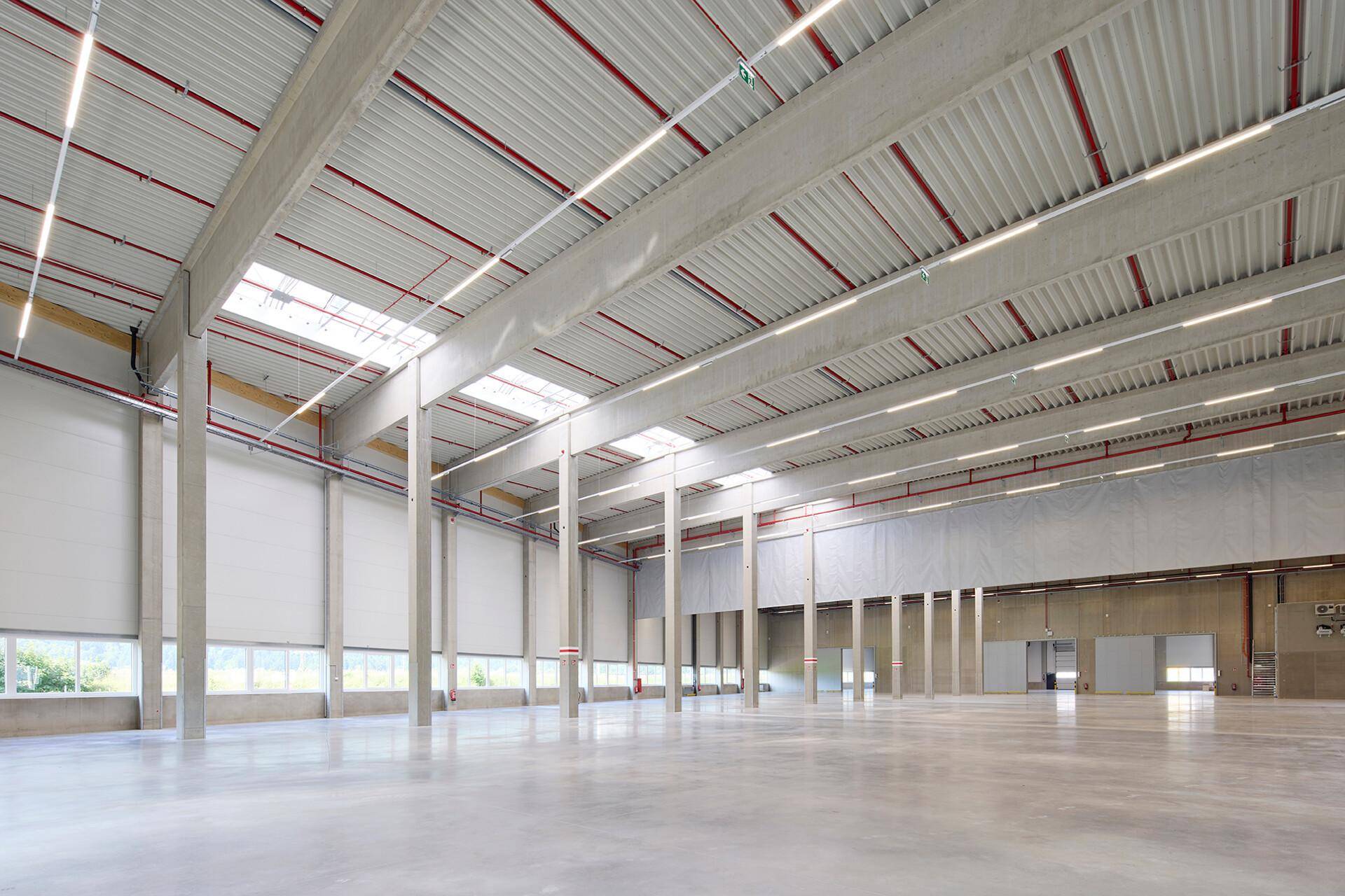 Geräumige, leere Lagerhalle mit hohen Decken, Betonpfeilern und natürlichem Lichteinfall durch Oberlichter.