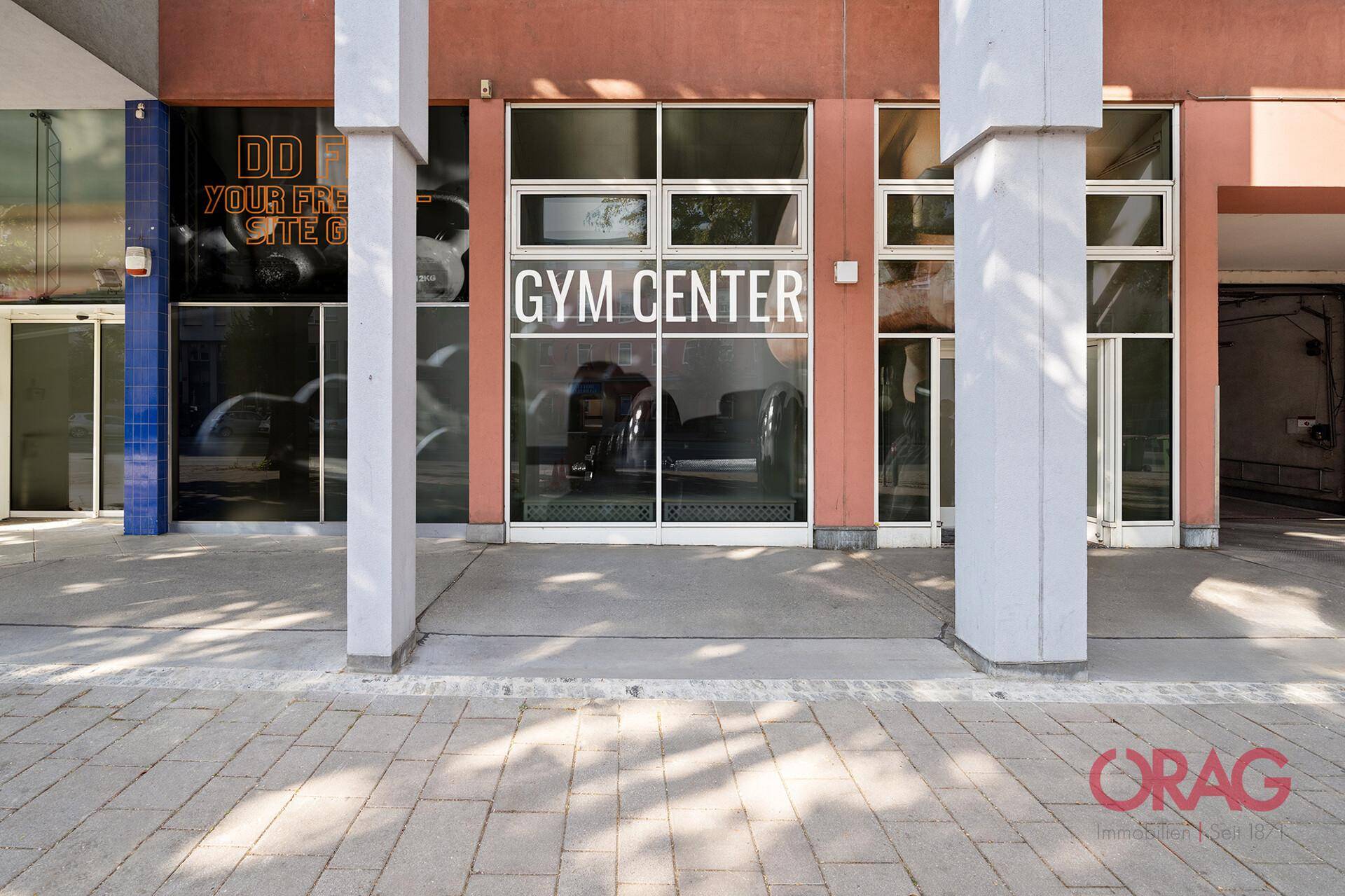 Eingangsbereich eines Gebäudes mit großen Glasfenstern und Beschilderung für ein Fitnesscenter.