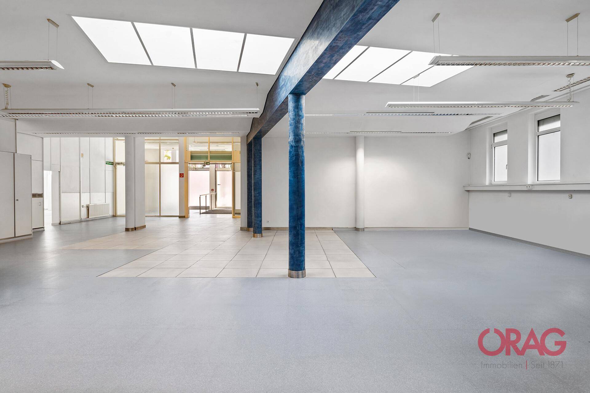 Geräumiger Gewerberaum mit Oberlichtern, blauen Säulen und verschiedenen Bodenbelägen, hell und offen.