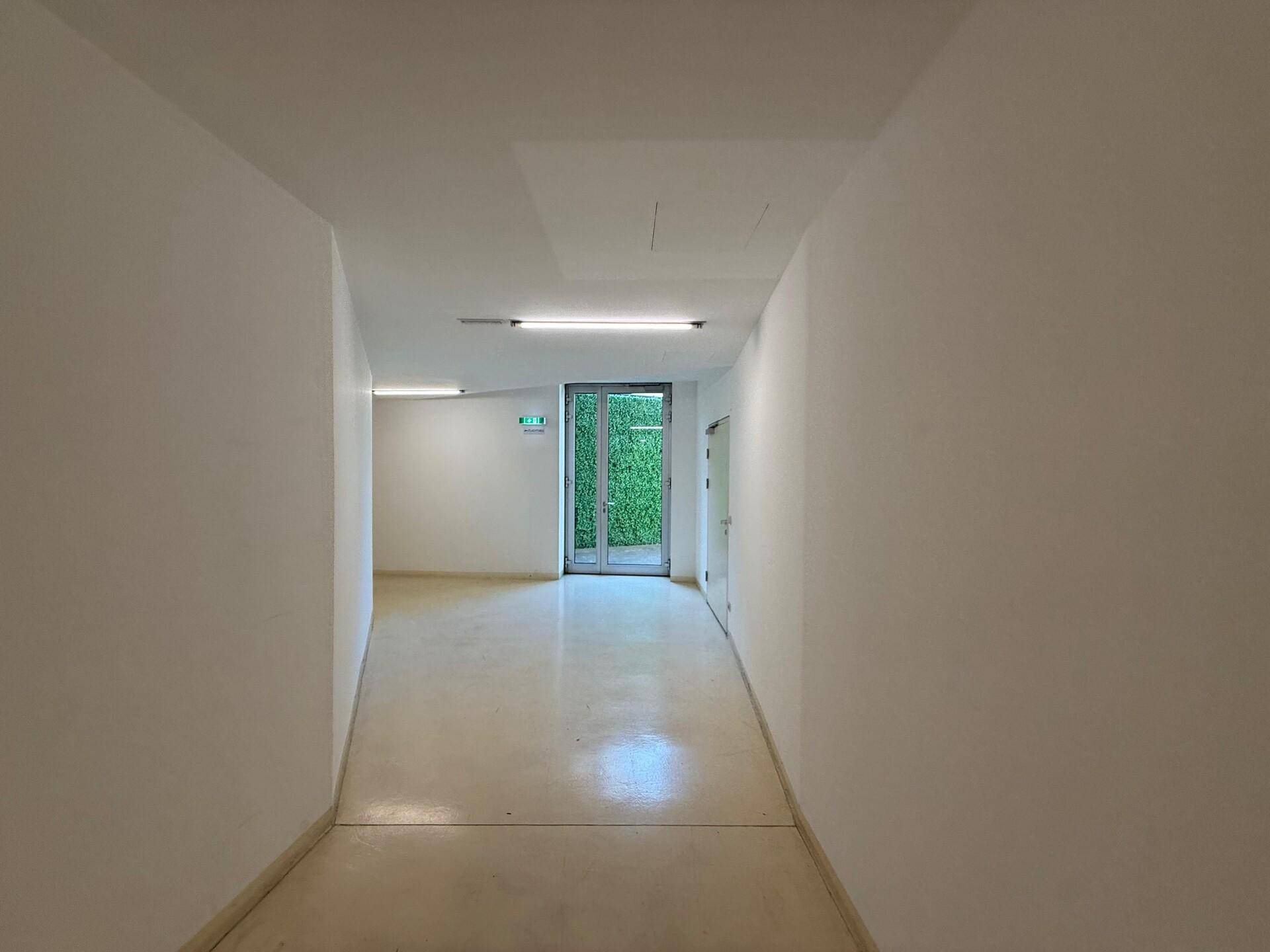 Langer, heller Flur mit weißen Wänden und hellem Boden, der zu einem Ausgang mit Grünfläche führt.