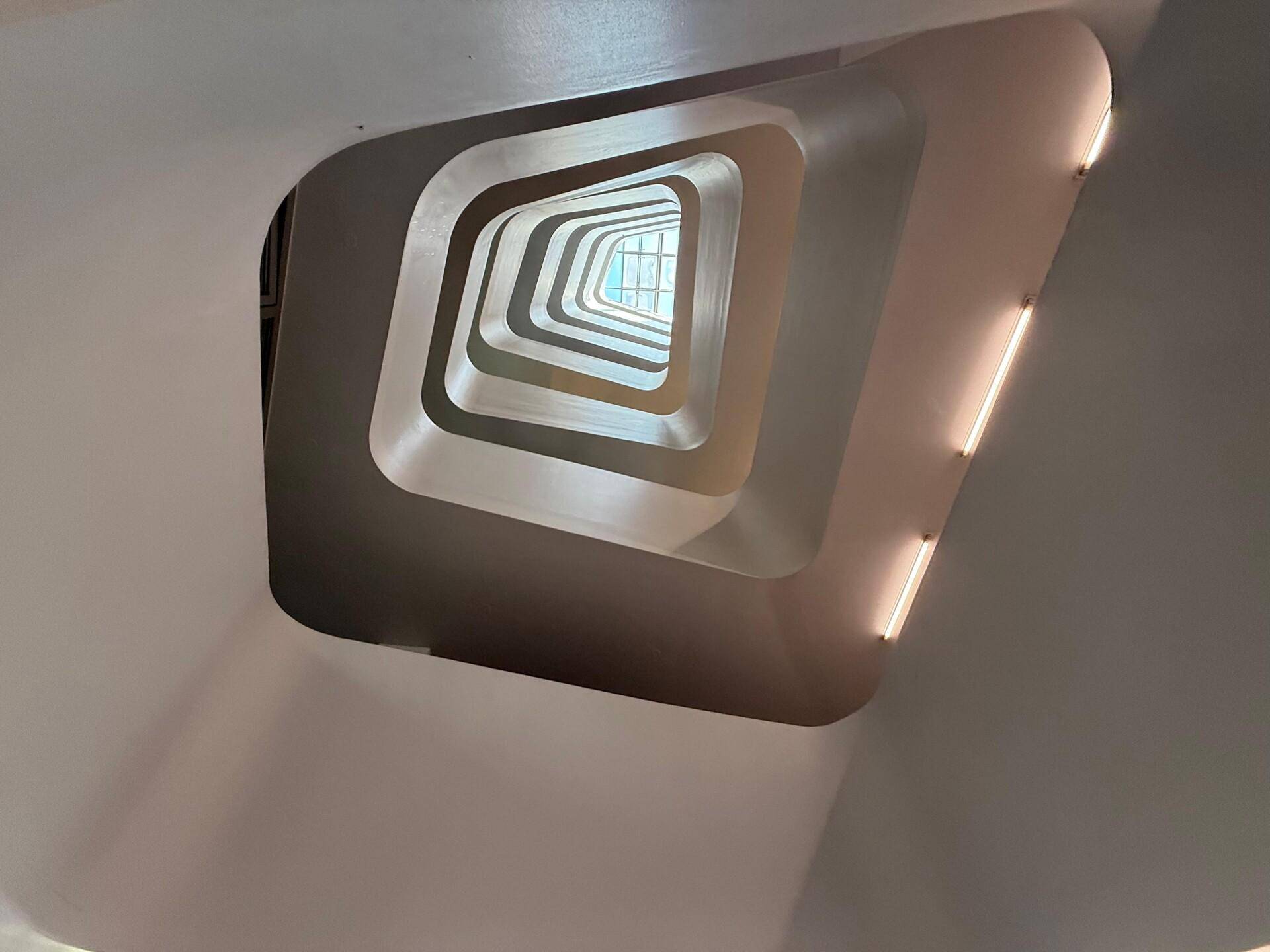 Blick nach oben in ein modernes, geometrisches Treppenhaus, das durch seine Formgebung beeindruckt und viel Licht hereinlässt.