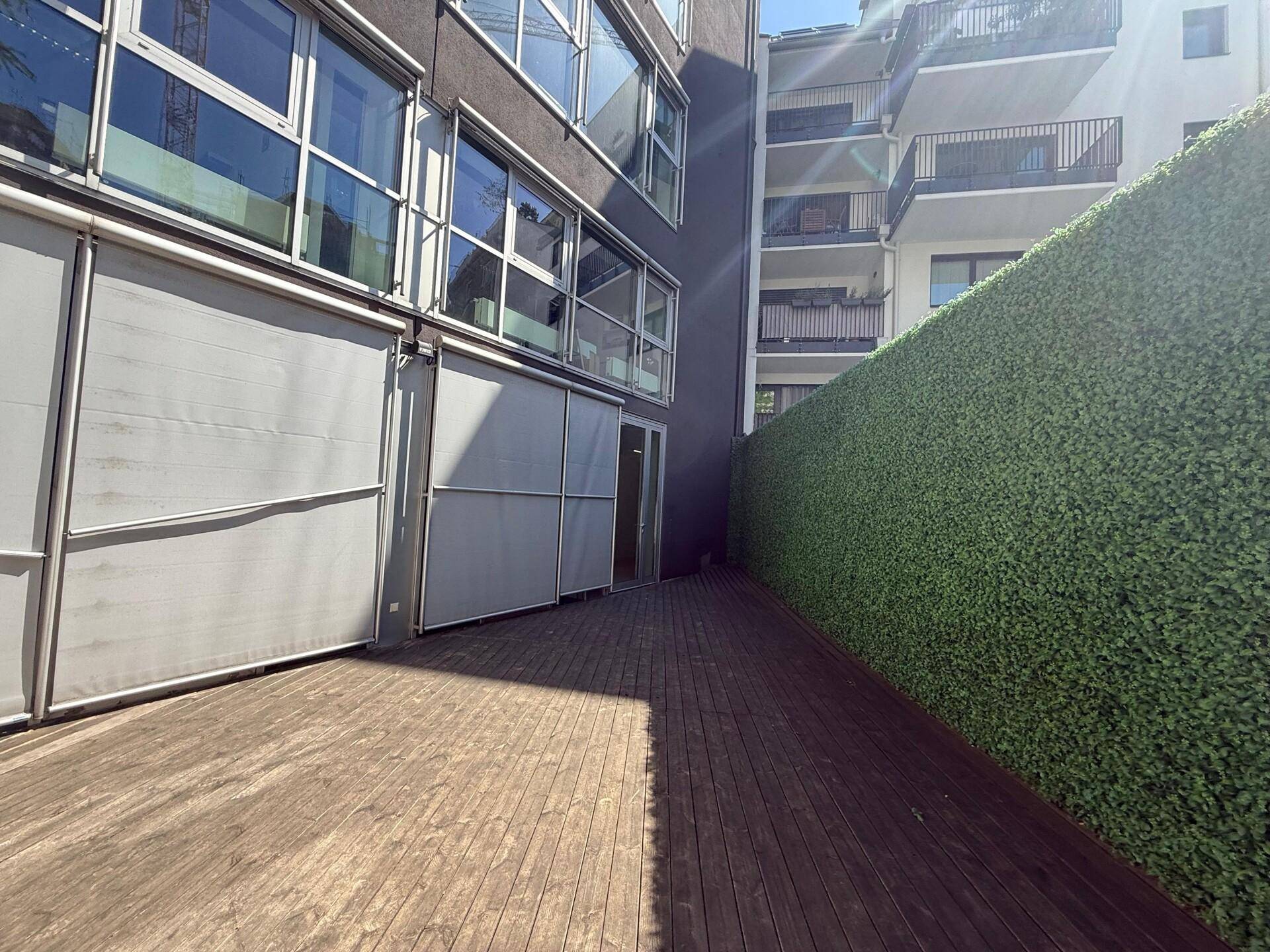 Sonniger Außenbereich mit Holzterrasse, einer hohen grünen Hecke und der modernen Gebäudefassade.