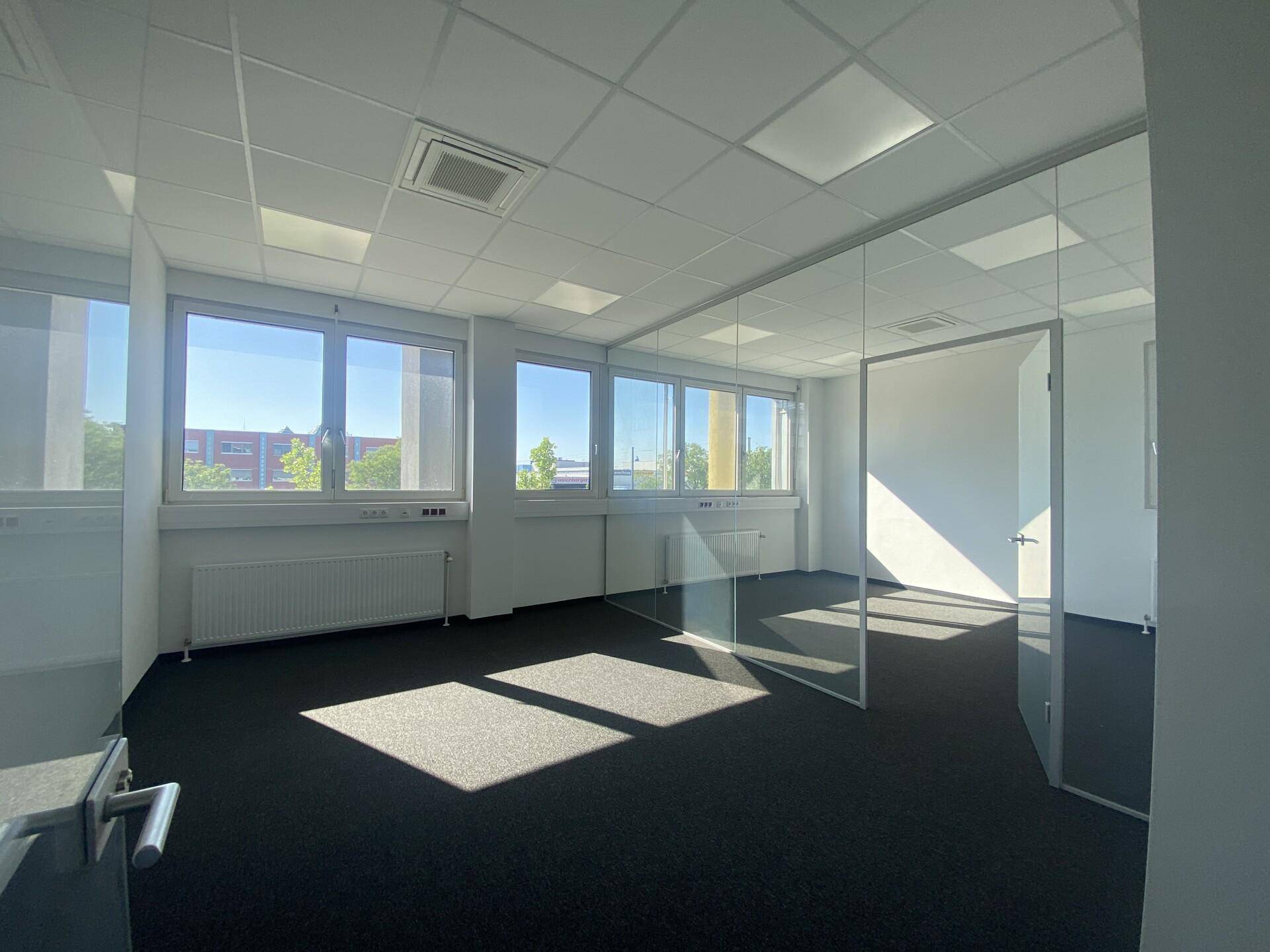 Helles Büro mit Teppichboden, Glaswänden und einer Glastür, durch die Sonnenlicht in den Raum fällt.
