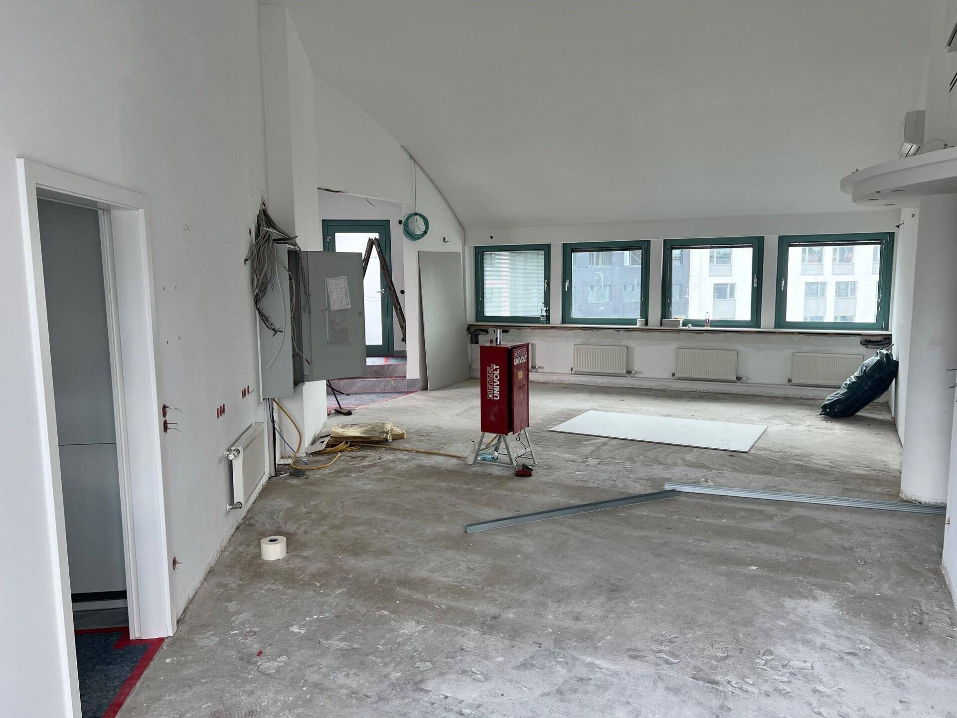 Großer Raum im Rohbauzustand mit Betonboden, weißen Wänden und einer Fensterreihe, bereit für den Ausbau.