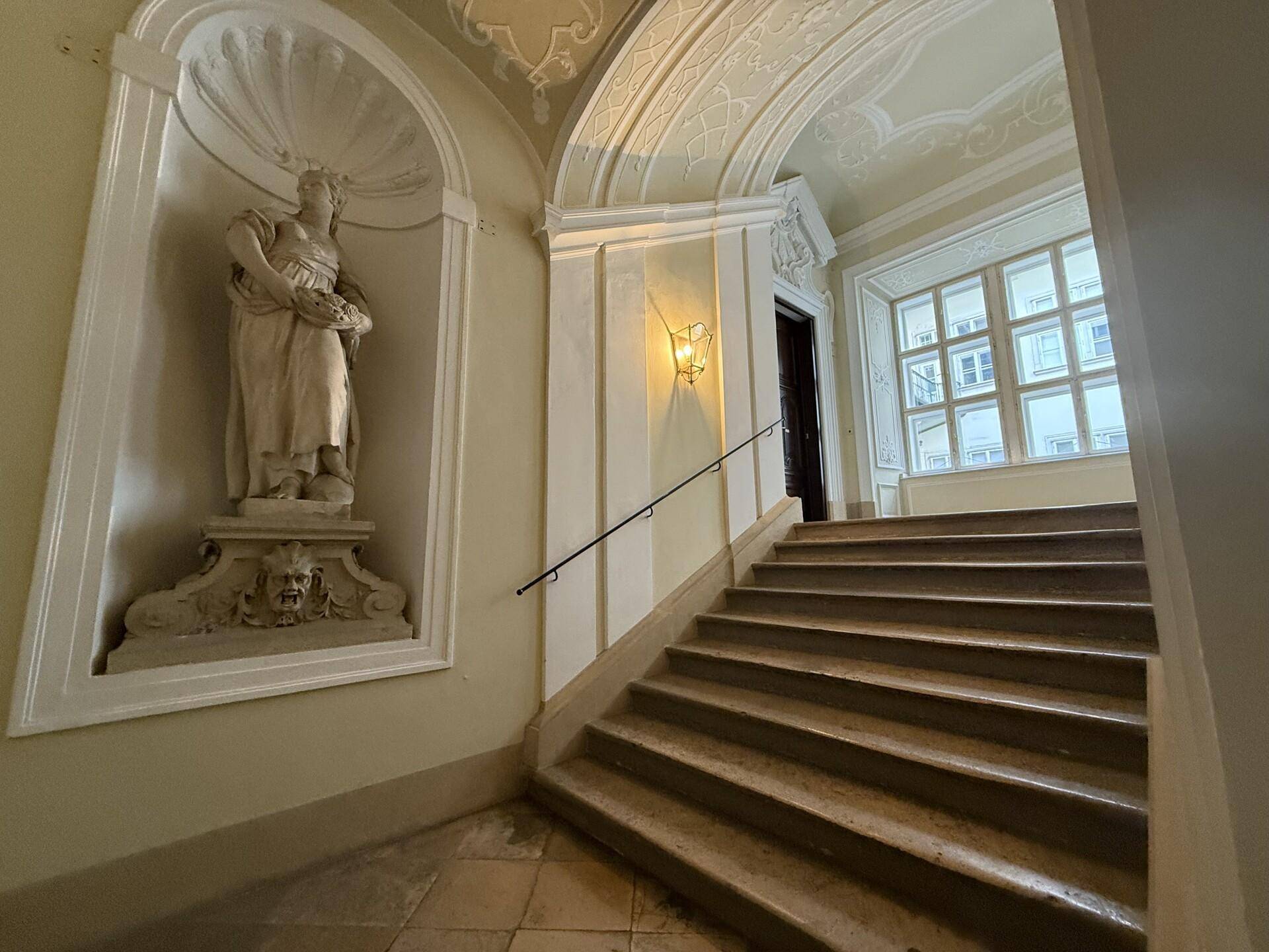 Historisches Treppenhaus mit einer Statue in einer Nische, verzierten Decken und großen Fenstern.