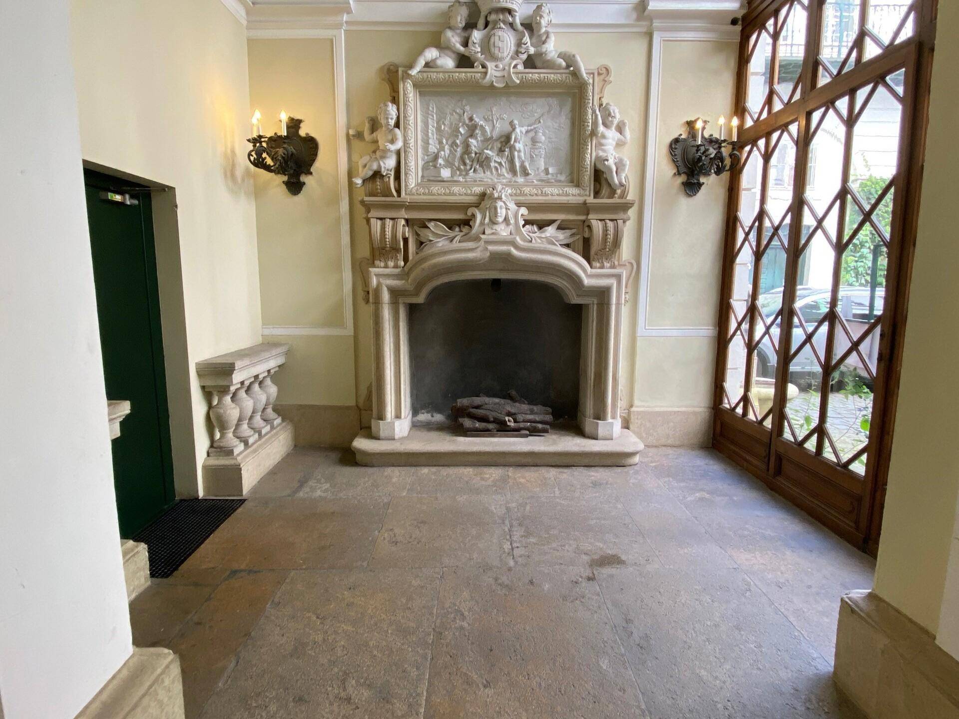 Elegantes Foyer mit einem großen, verzierten Kamin, Steinboden und hohen Holztüren zu einem Außenbereich.