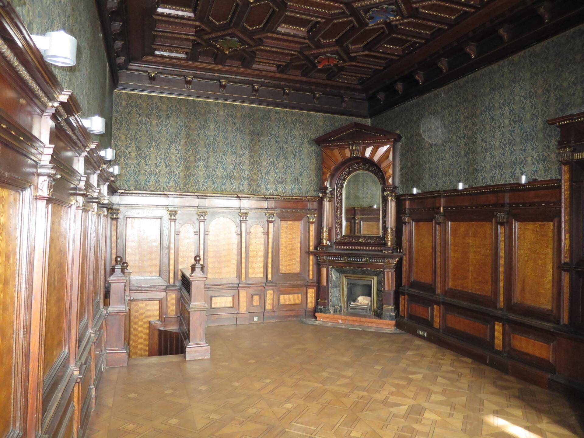 Historischer Raum mit dunkler Holzvertäfelung, gemusterter Tapete, Kassettendecke und einem Kamin.
