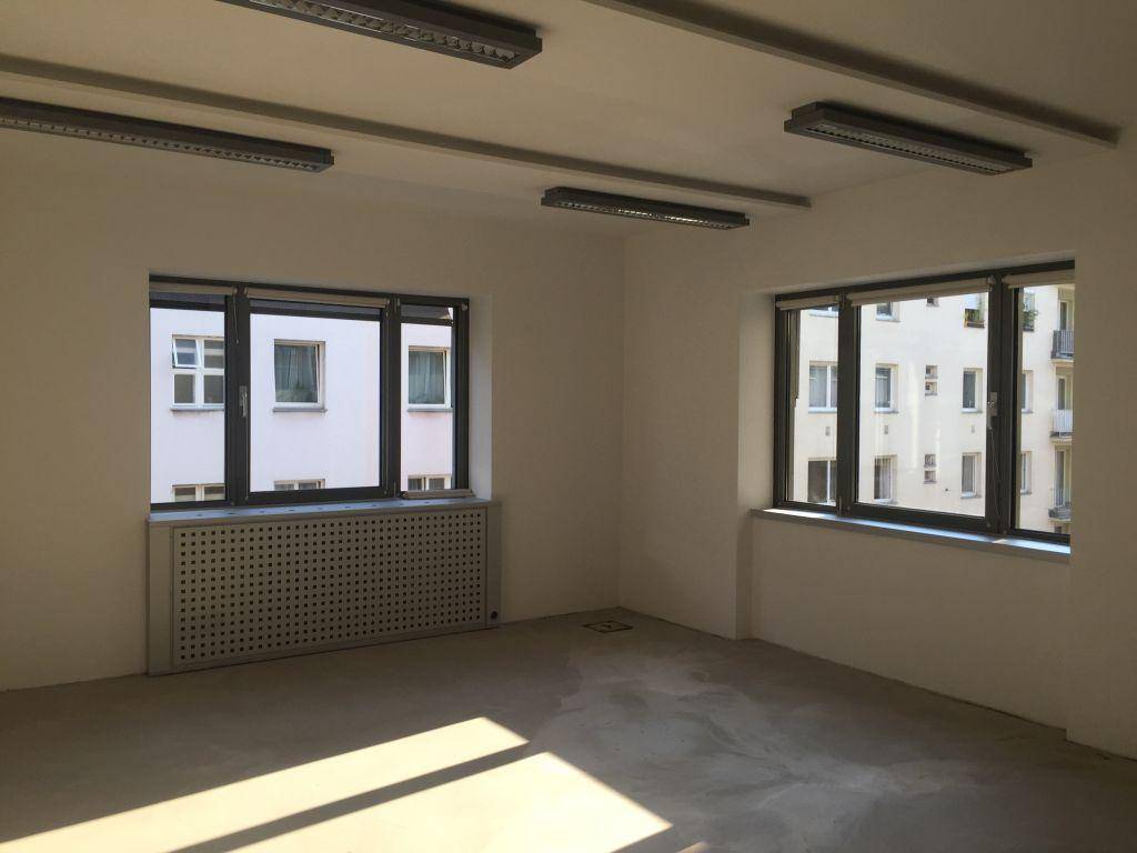 Leeres Büro mit weißen Wänden, großen Fenstern und Blick auf gegenüberliegende Gebäude.