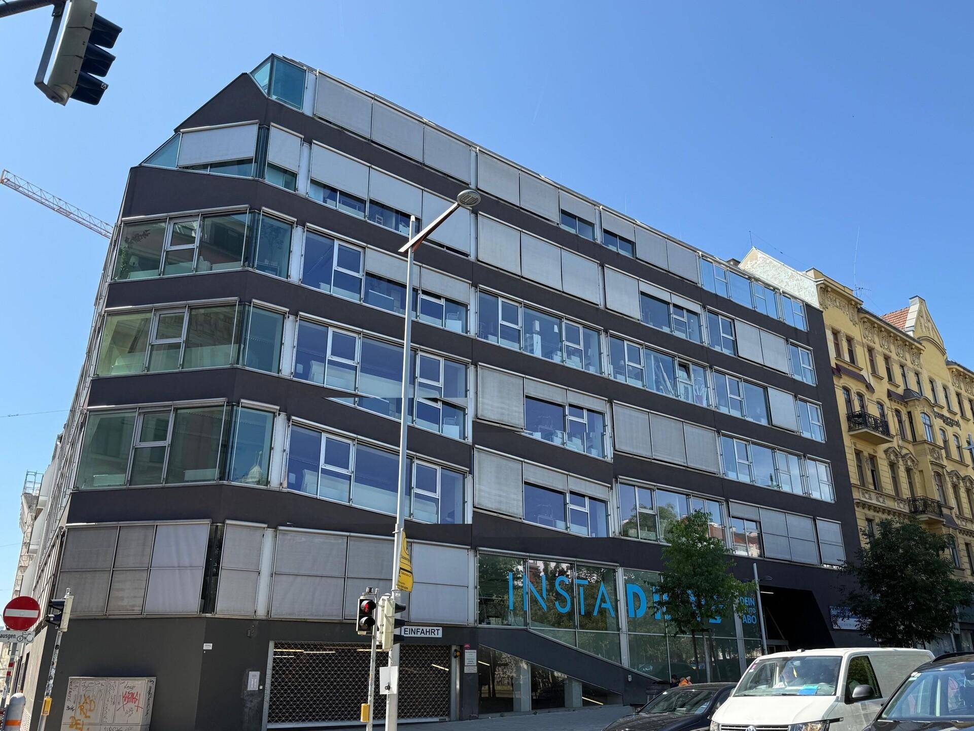 Modernes Bürogebäude mit großen Fenstern und Jalousien an einer städtischen Straße unter blauem Himmel.