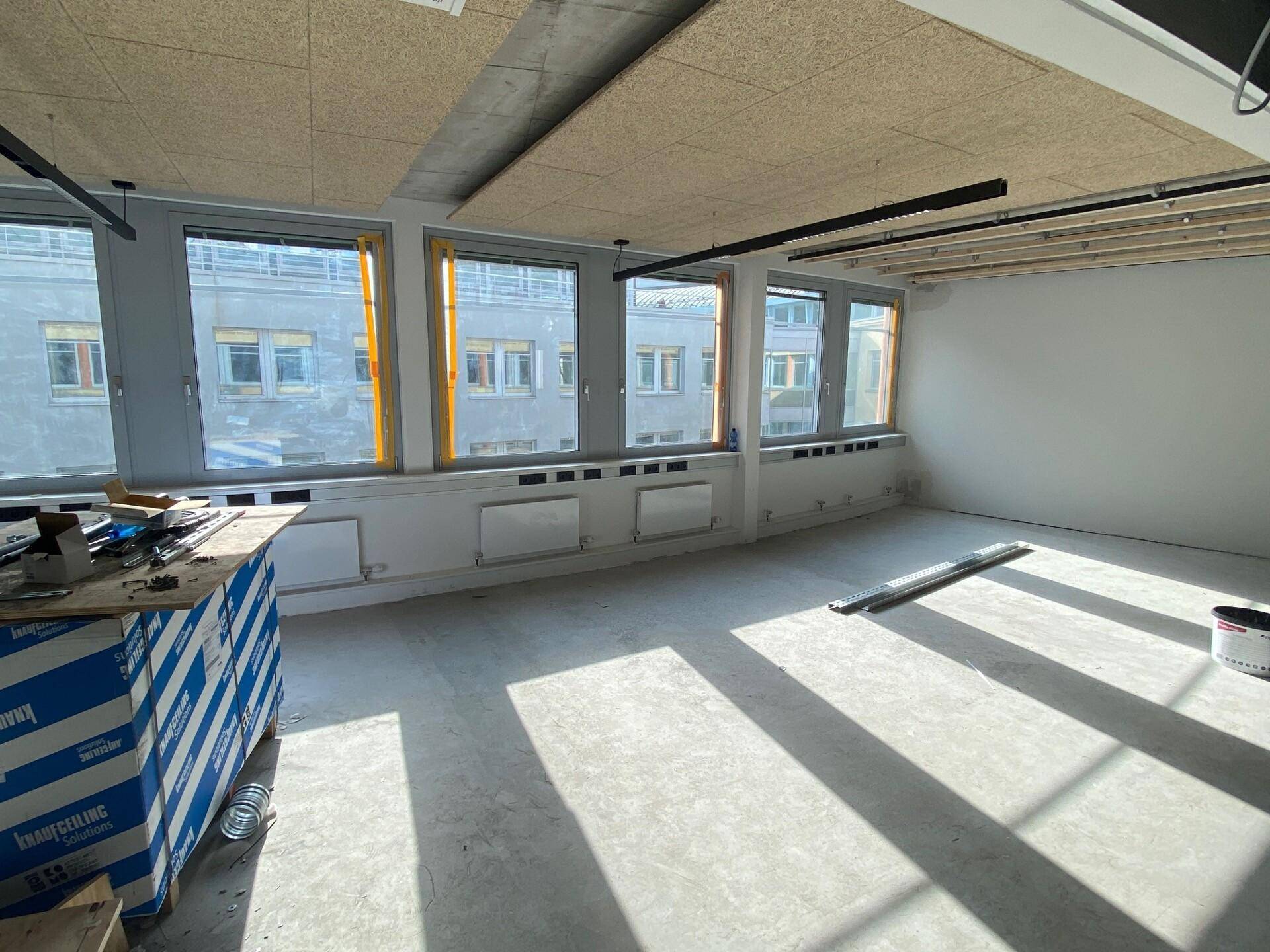 Unmöblierter Büroraum im Rohbau mit Betonboden, Akustikdecke und großen Fenstern.