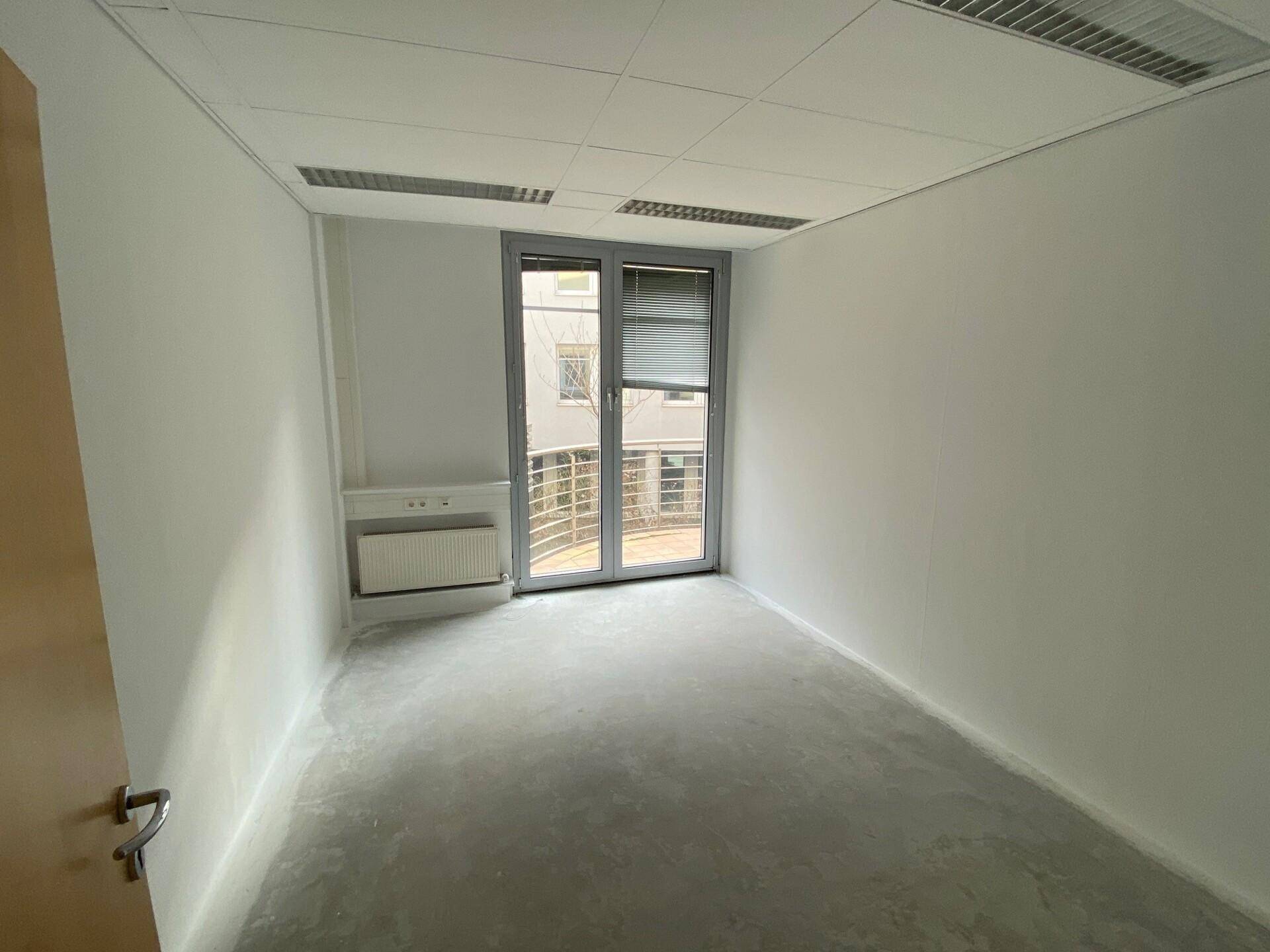 Leerer Büroraum mit Betonboden, weißen Wänden und Zugang zu einem Balkon oder einer Terrasse.