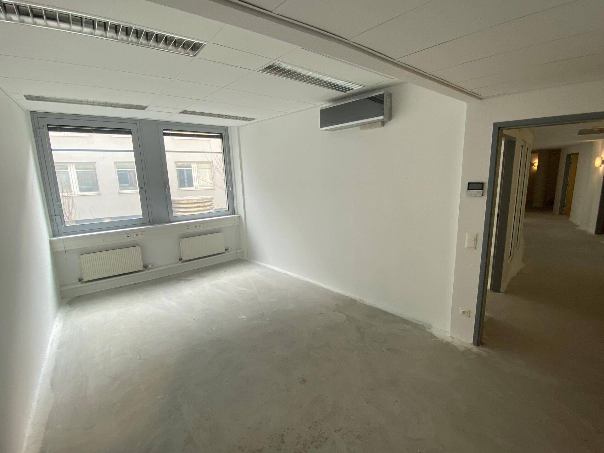 Leerer Büroraum mit Betonboden, weißen Wänden, Rasterdecke und Klimaanlage.
