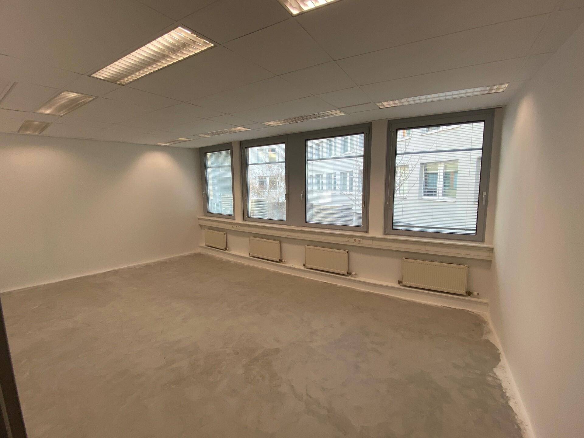 Leerer Büroraum mit Betonboden, weißen Wänden, Rasterdecke und Fenstern mit Stadtblick.