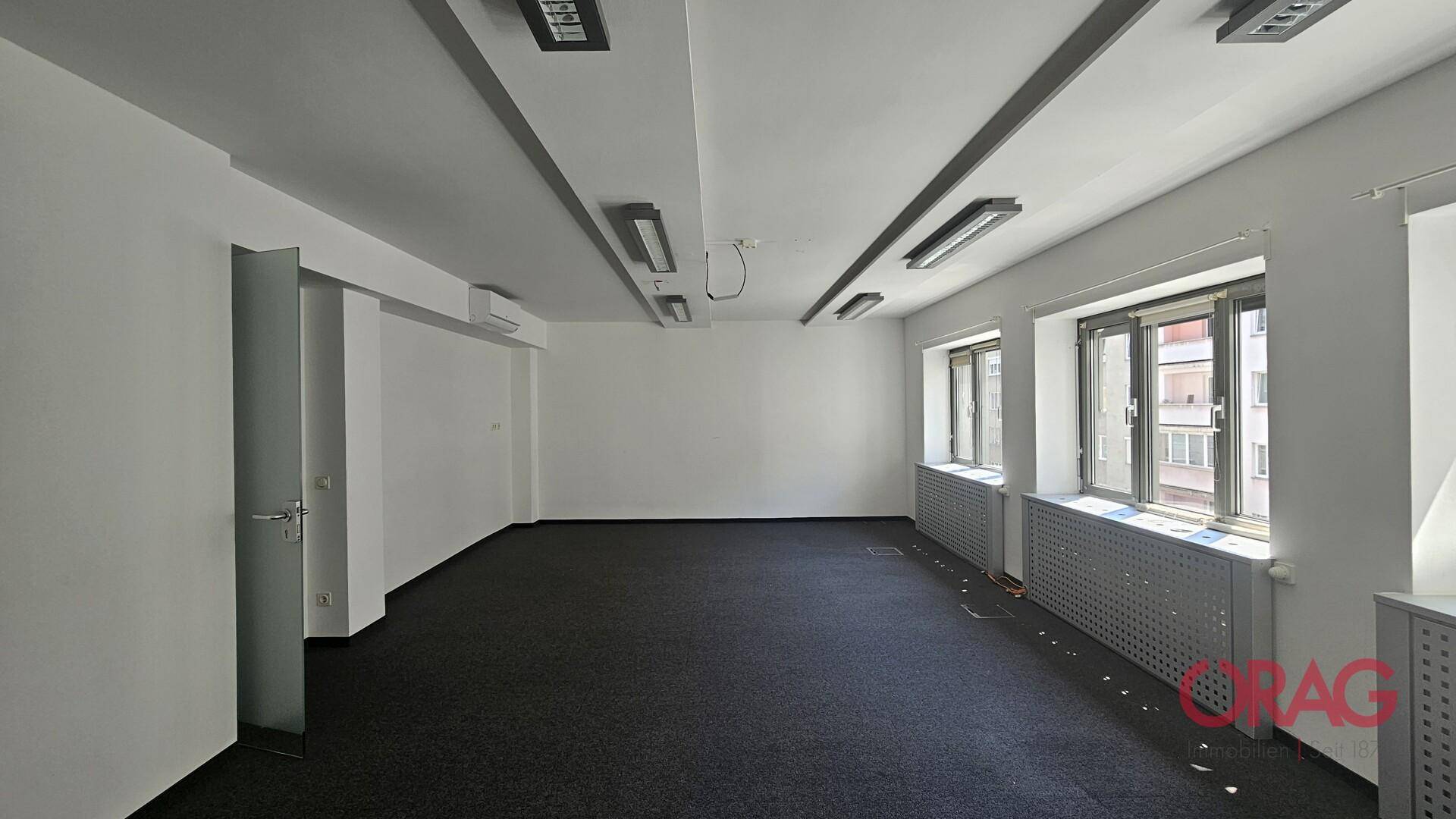 Geräumiges, leeres Büro mit Teppichboden, großen Fenstern und Klimaanlage, bereit zur individuellen Gestaltung.
