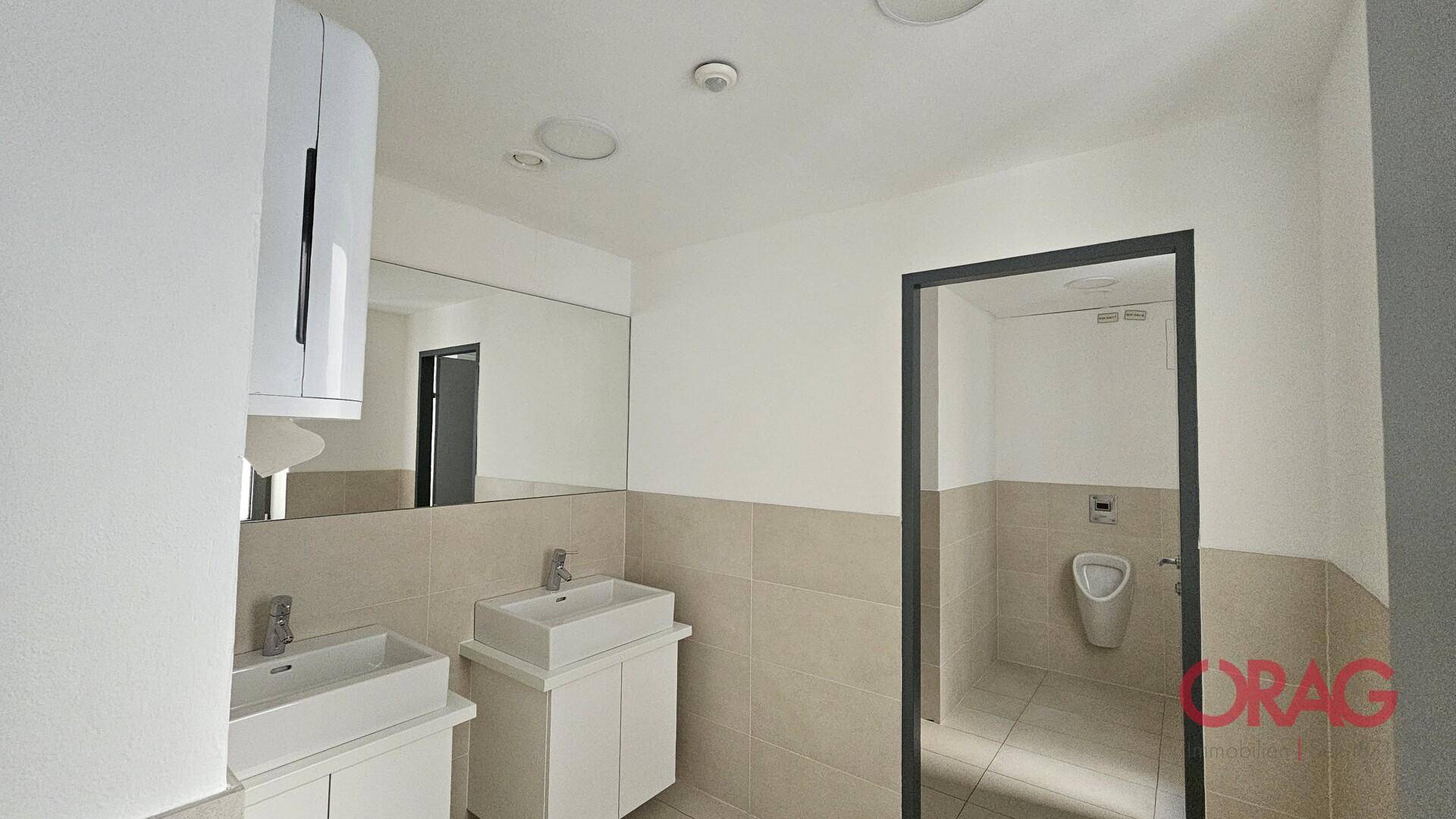 Modernes Badezimmer mit zwei Waschbecken, großem Spiegel, hellen Fliesen und einem Urinal.