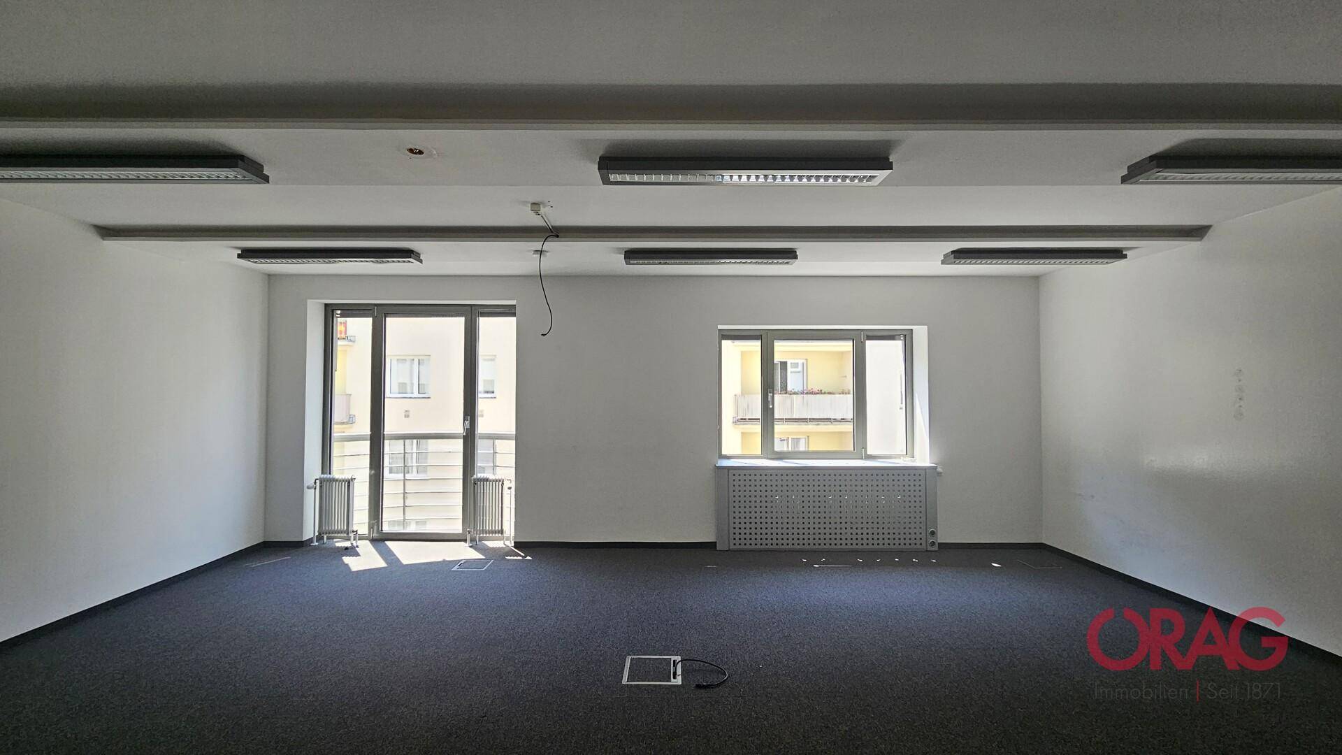 Heller Büroraum mit Teppichboden, großen Fenstern und einer Balkontür, die viel Tageslicht hereinlässt.