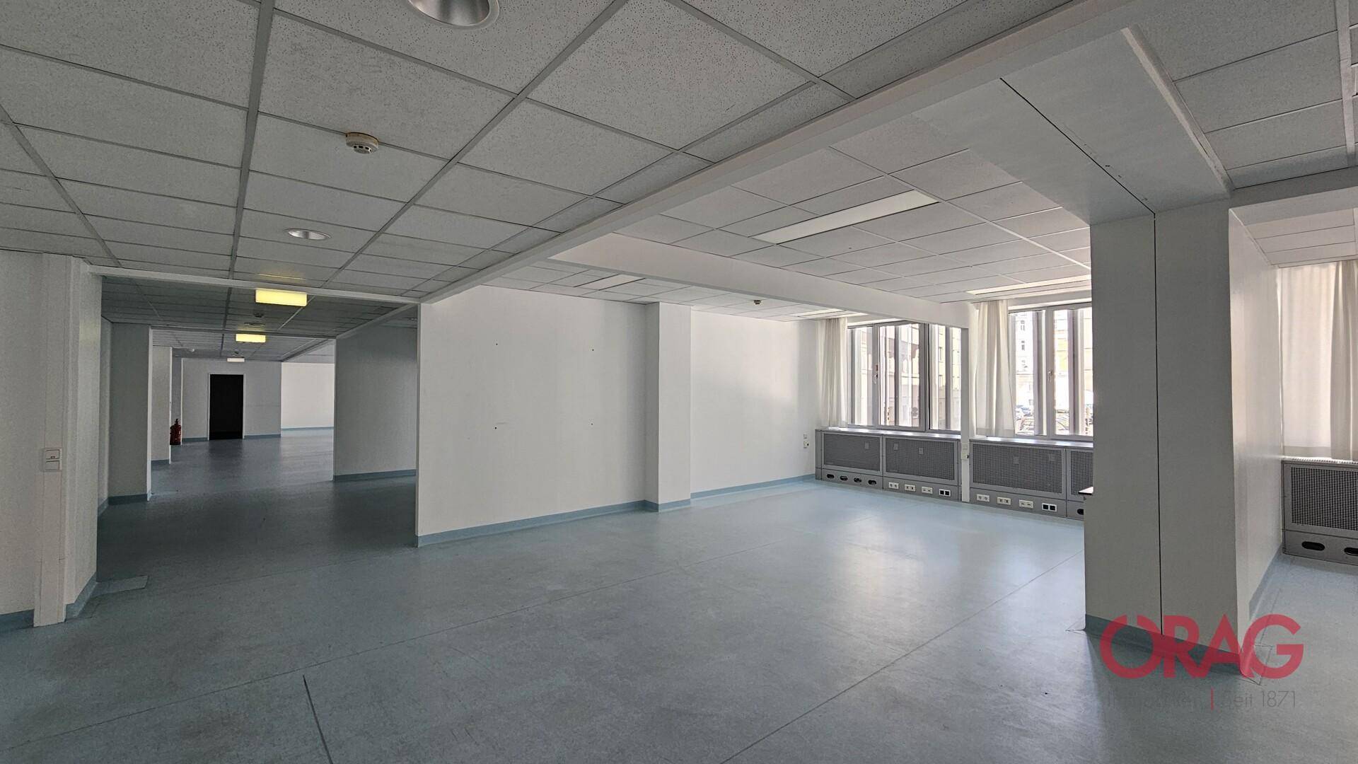 Heller Büroraum mit großen Fenstern, Teppichfliesen und abgehängter Decke.