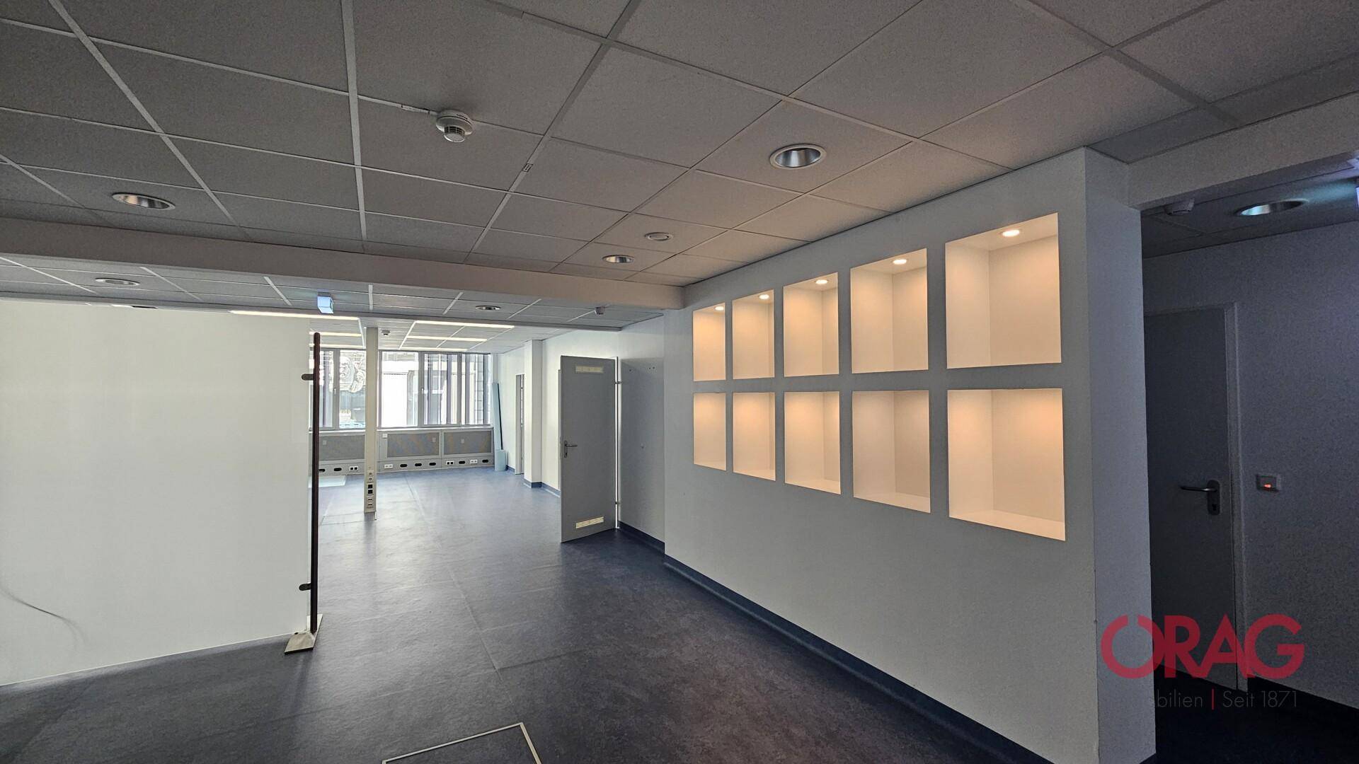 Geräumiger Bürobereich mit beleuchteten Einbauregalen, abgehängter Decke und Fenstern im Hintergrund.