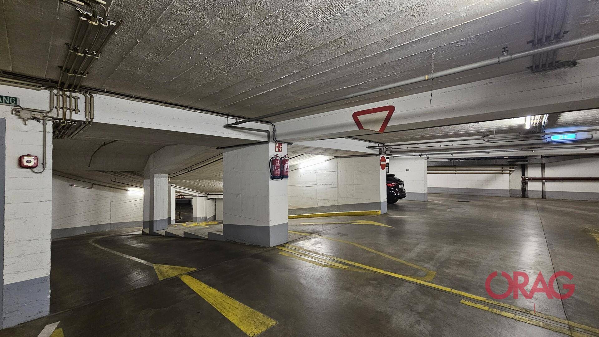 Tiefgarage mit Betonboden, sichtbaren Leitungen und gelben Fahrbahnmarkierungen für Fahrzeuge.