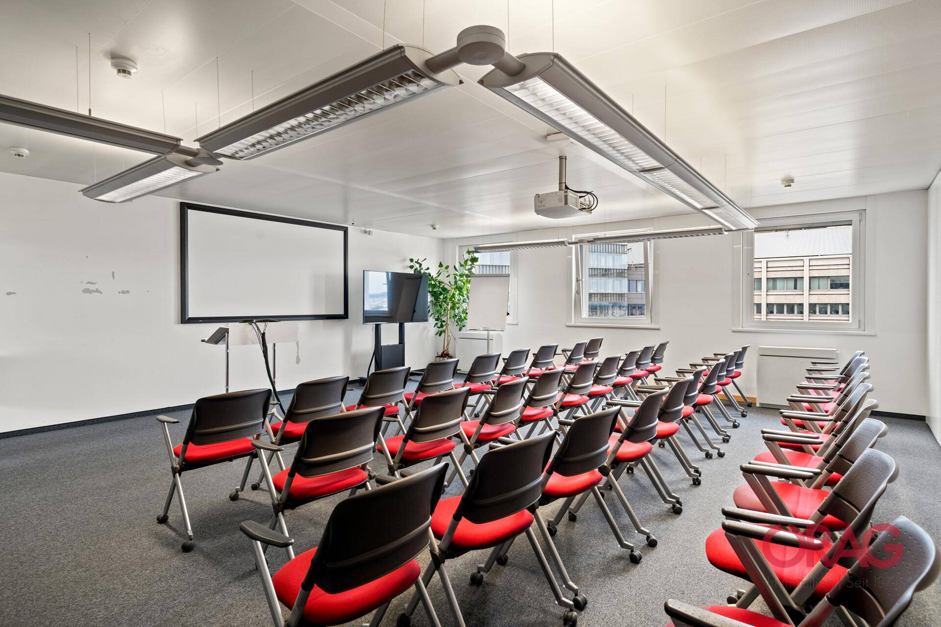 Großer Konferenzraum mit Reihen von roten Stühlen, Projektionsfläche und Tageslicht.