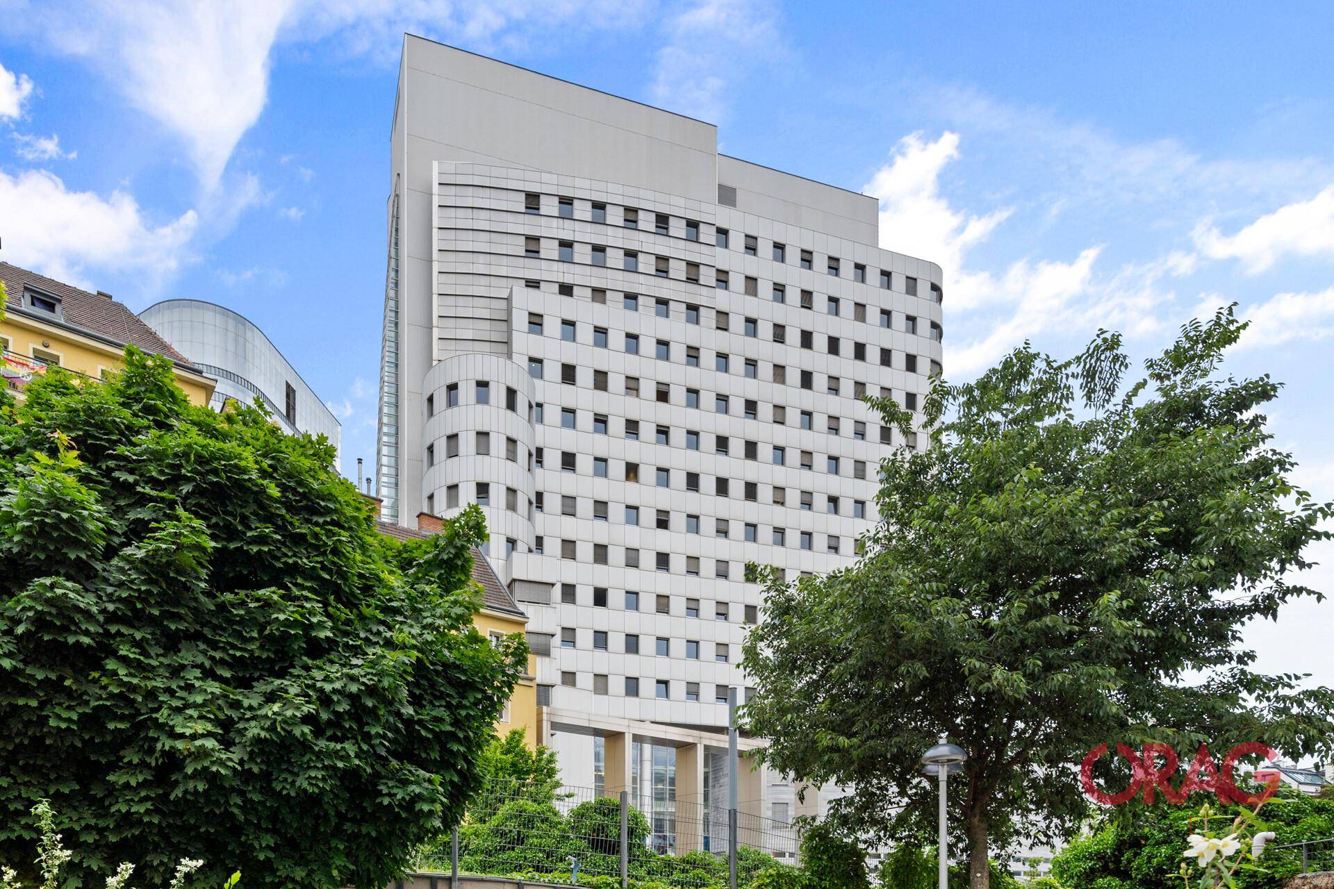 Modernes Hochhaus mit vielen Fenstern, umgeben von grünen Bäumen unter blauem Himmel.