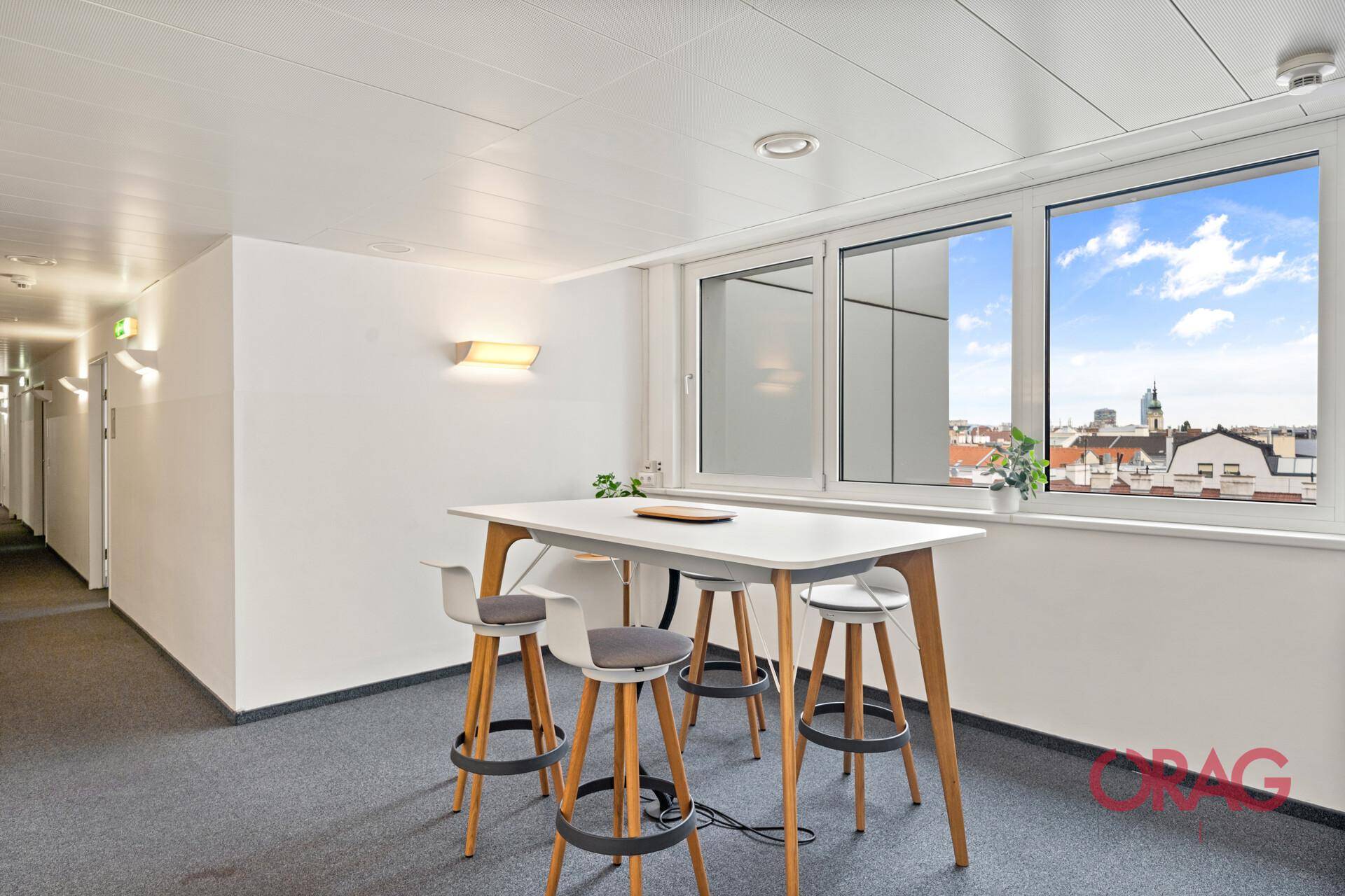 Heller Loungebereich mit Stehtisch, Hockern und Fenstern mit Stadtblick.