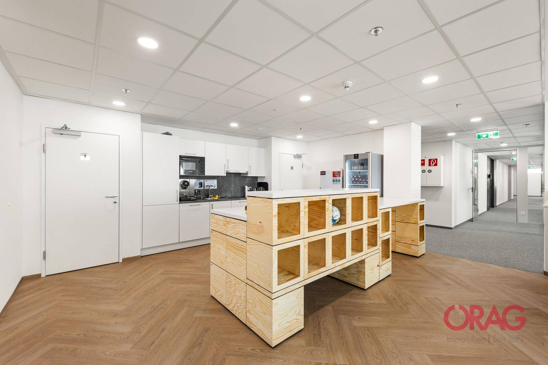 Moderne Büroküche mit weißen Schränken, Geräten und Fischgrätparkett, ideal für Pausen.