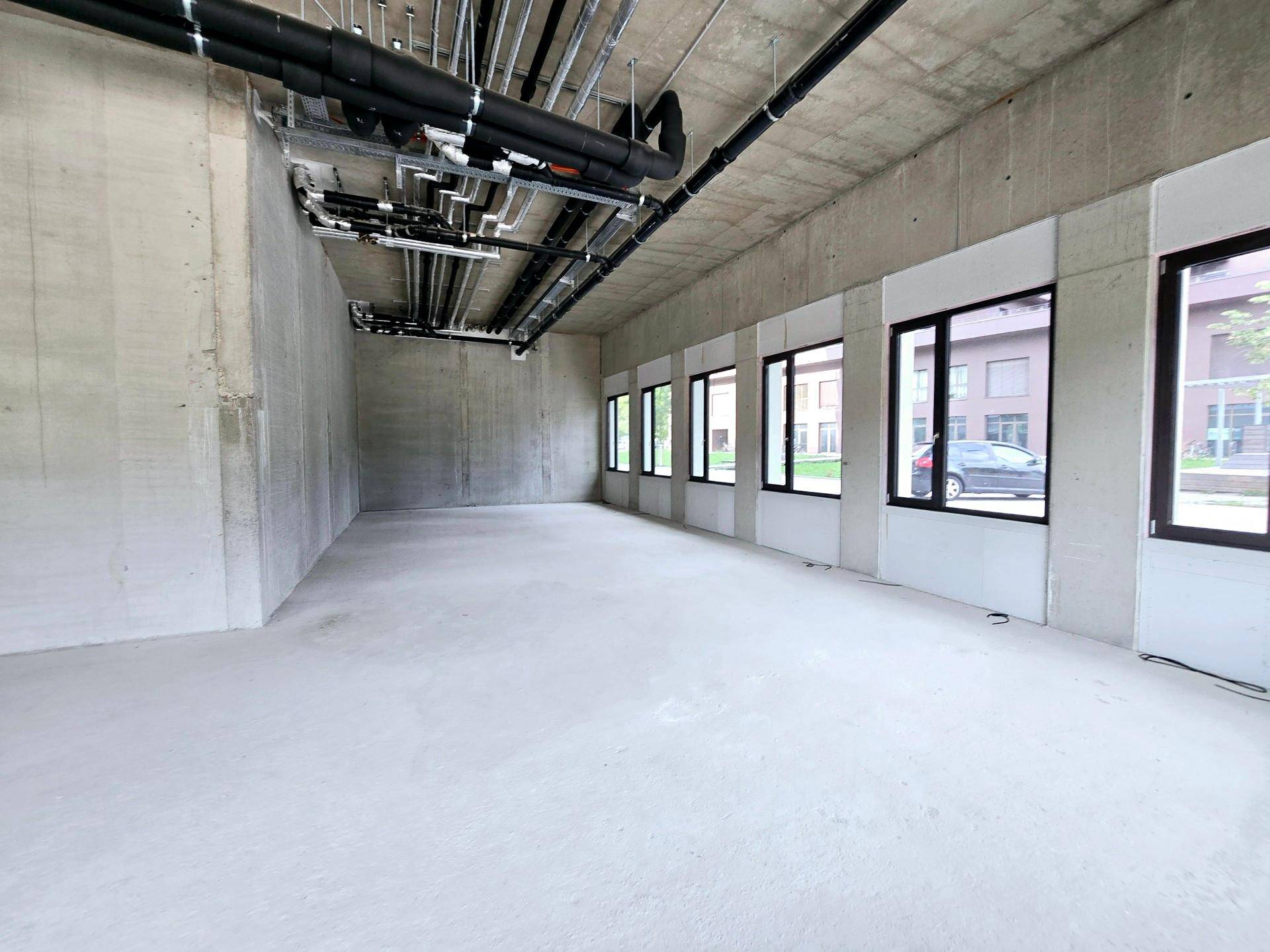 Unfertige Gewerbeeinheit mit Betonboden, freiliegenden Rohren und mehreren Fenstern.