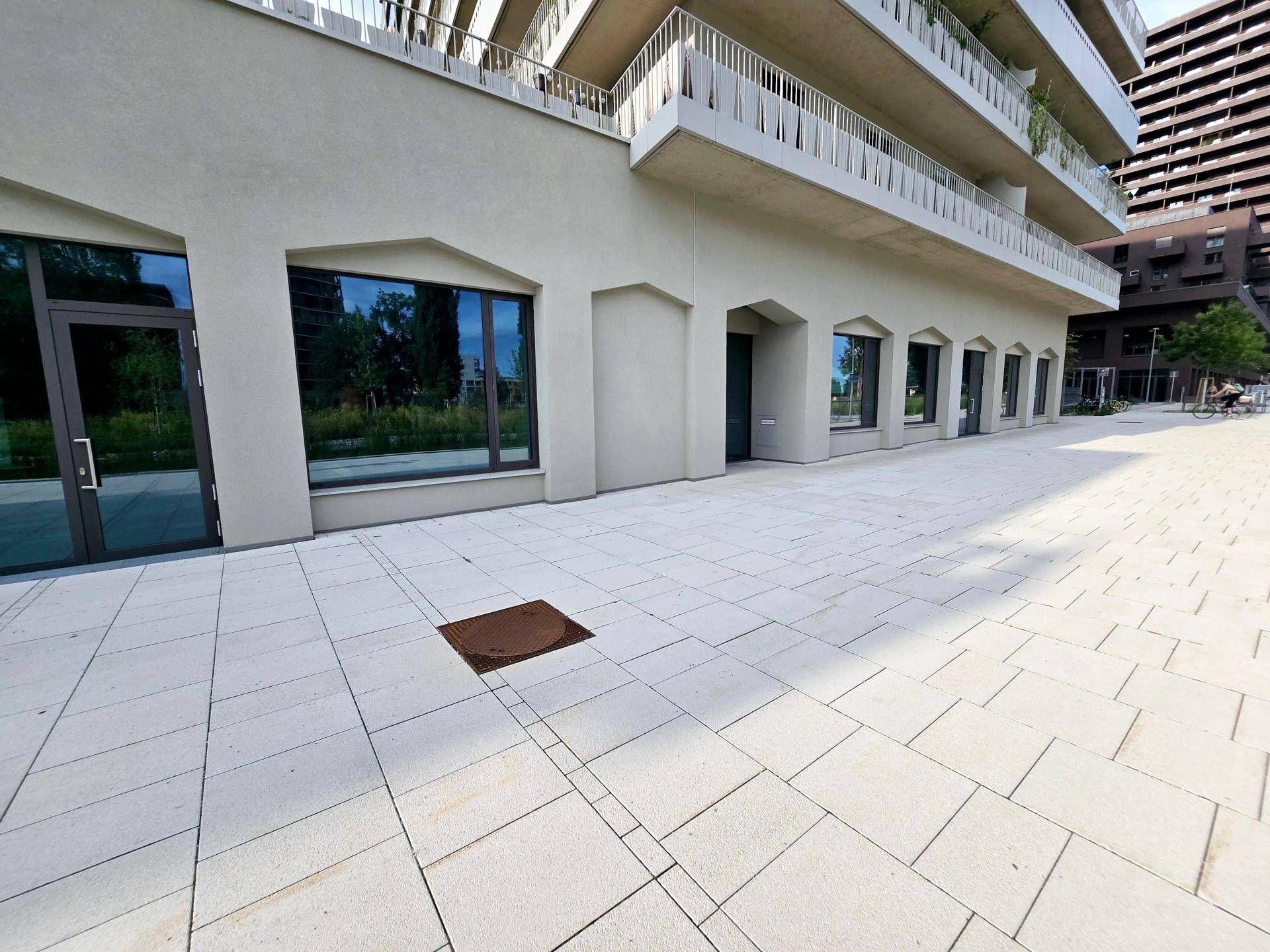Moderne Gebäudefassade mit ebenerdigen Gewerbeeinheiten und einem breiten, gefliesten Vorplatz.