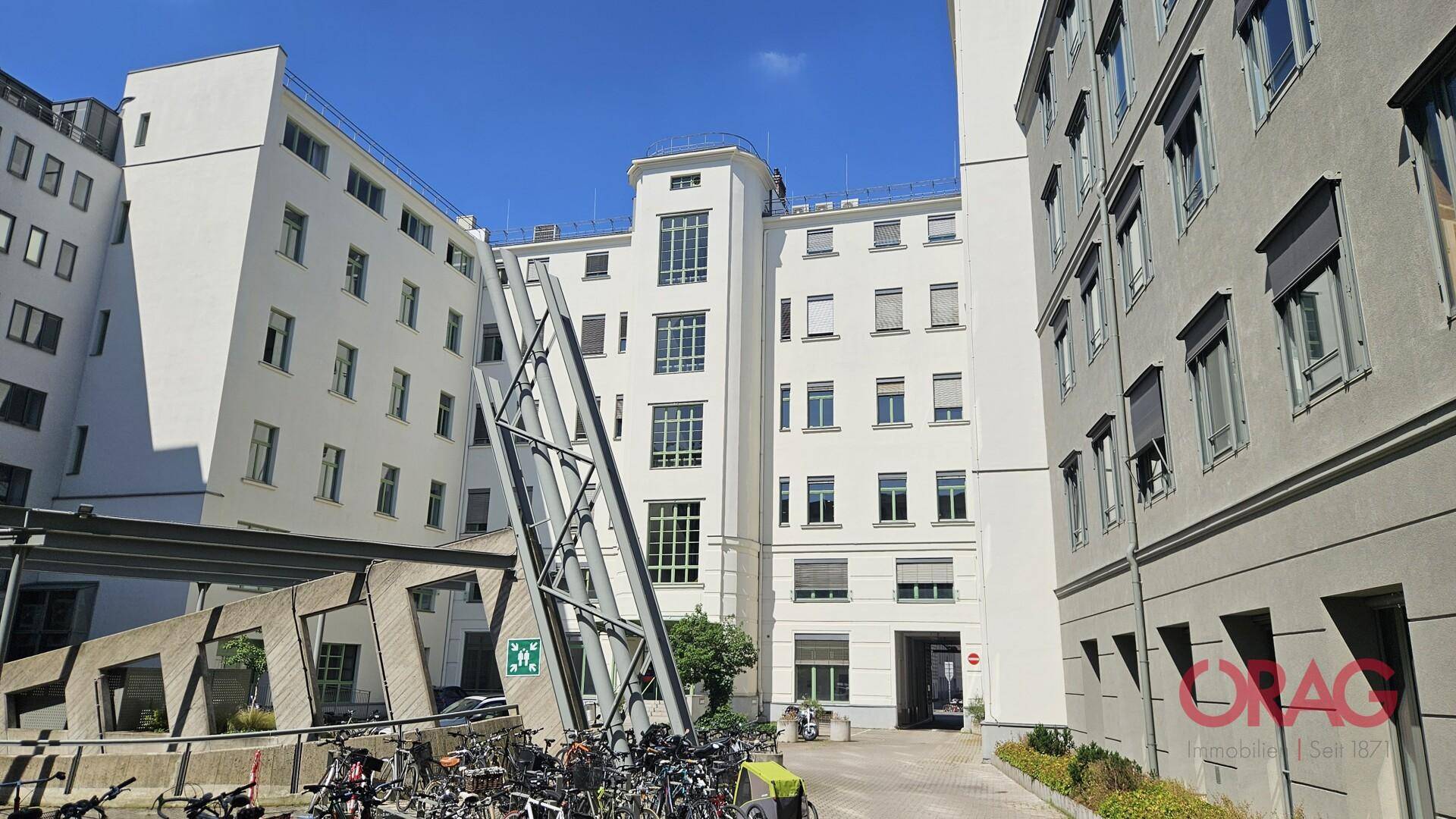 Innenhofansicht eines modernen Bürokomplexes mit Fahrradständern und einem Durchgang zu weiteren Gebäuden.