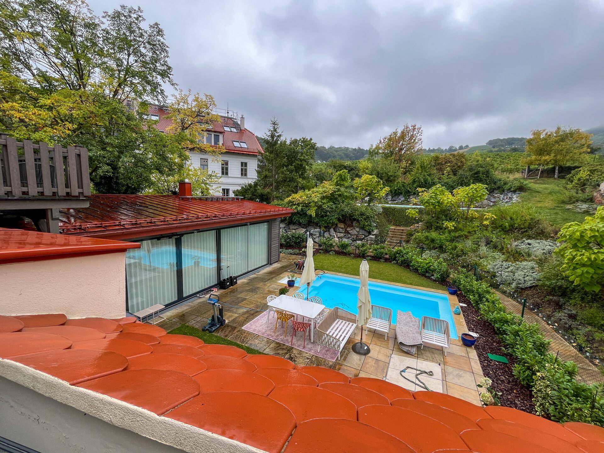Panoramablick auf den Garten mit Pool, Terrasse und umliegender grüner Landschaft unter bewölktem Himmel.