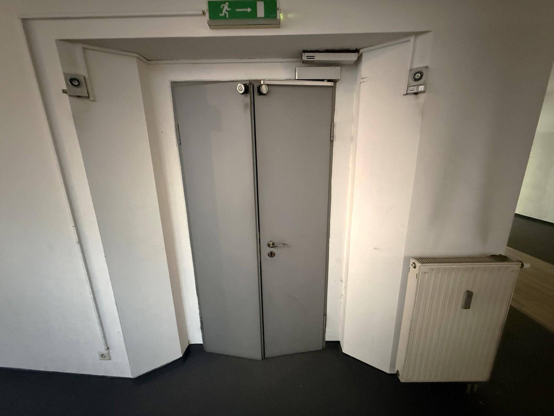 Doppeltür mit Notausgangsschild in einem schlichten Flur, daneben ein Heizkörper an der weißen Wand.