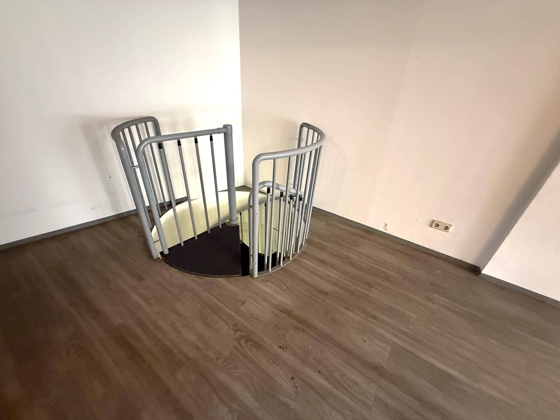 Kompakte Wendeltreppe mit Metallgeländer, die in einen unteren Bereich führt, umgeben von Laminatboden.