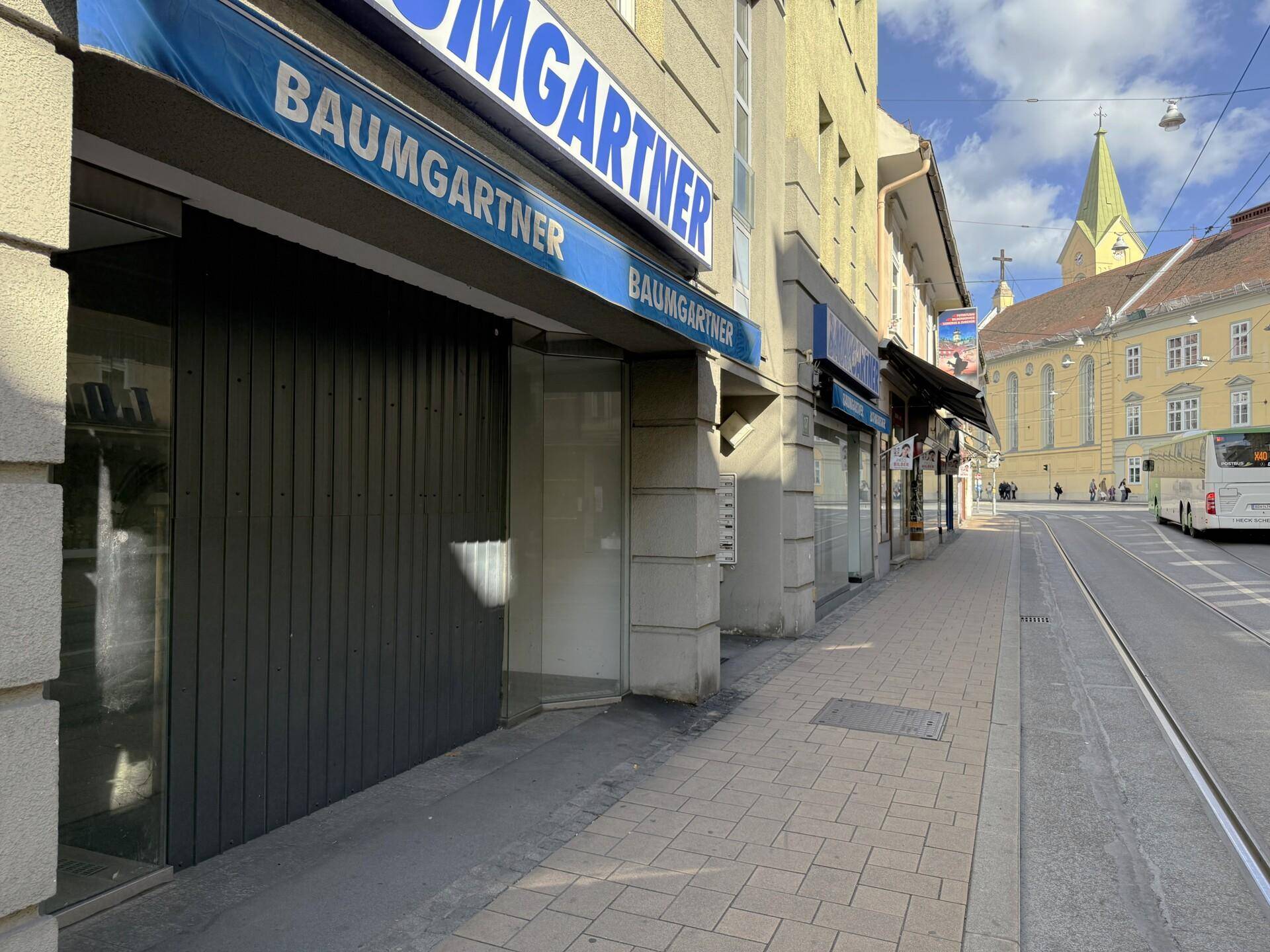 Städtische Straßenansicht eines Geschäftsgebäudes mit Markise und angrenzendem Bürgersteig, belebt durch Passanten und Verkehr.