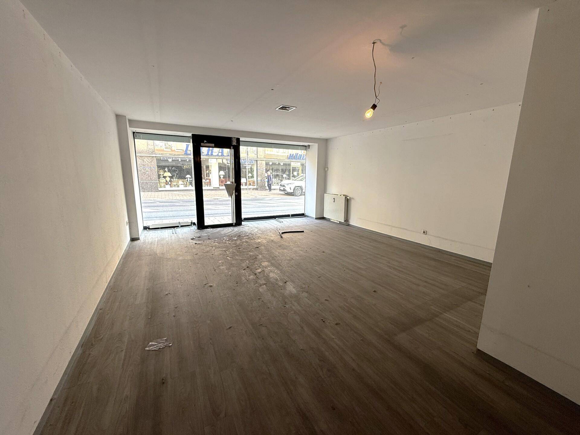 Leerer Ladenraum mit großen Schaufenstern zur Straße, Holzlaminatboden und weißen Wänden, bereit zur Einrichtung.