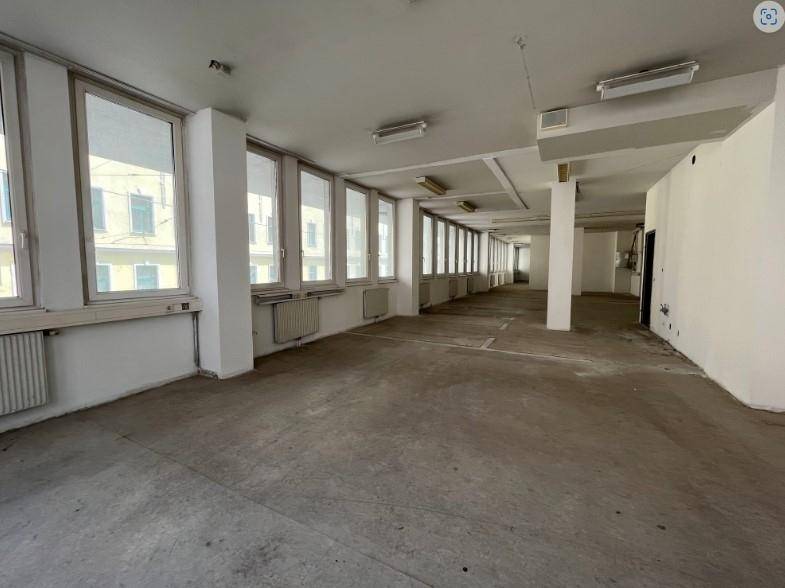 Großer, leerer Raum mit vielen Fenstern, Betonboden und Heizkörpern unter den Fenstern.