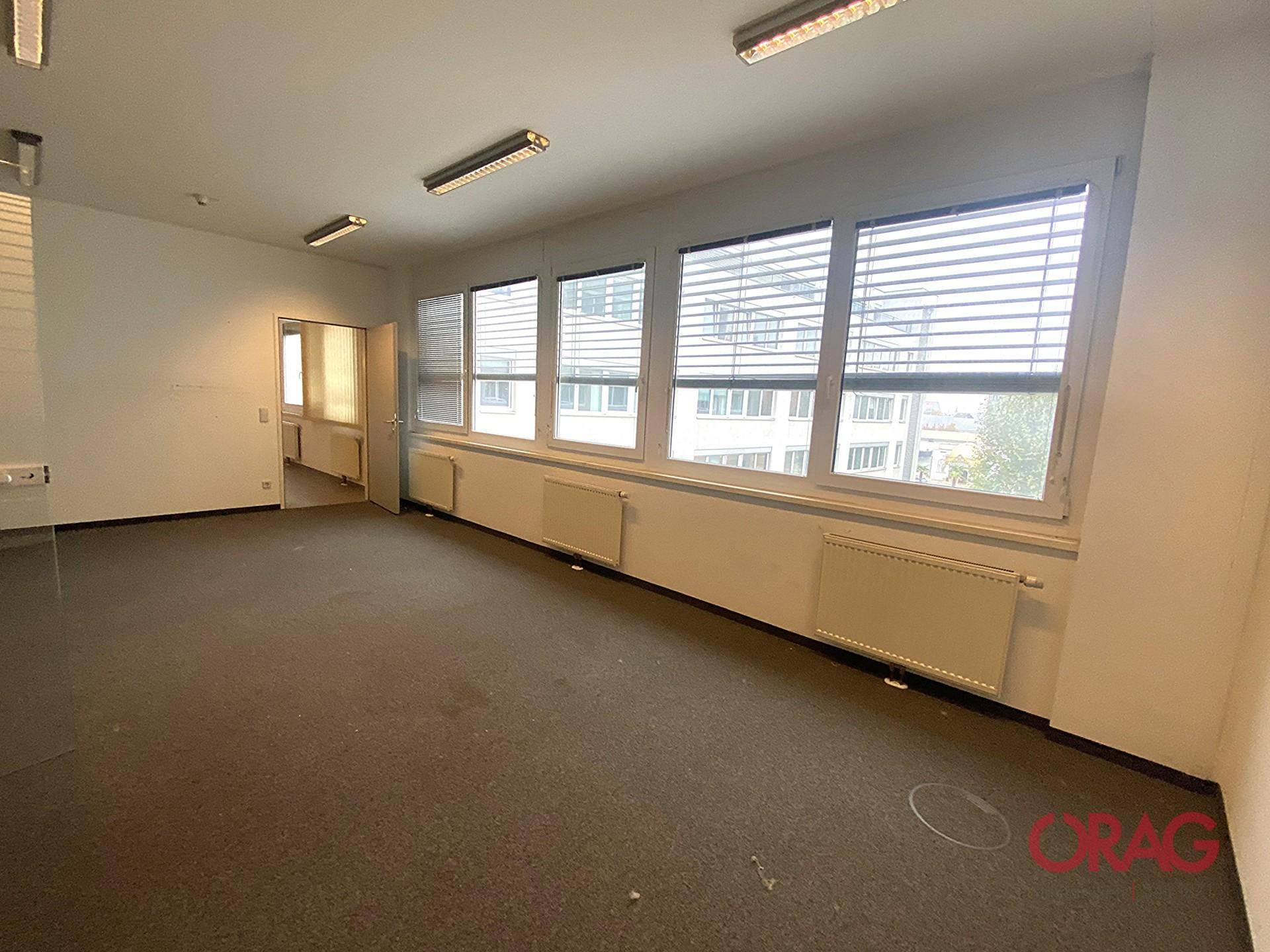 Heller Büroraum mit Teppichboden und Fensterfront, die einen Blick auf die umliegenden Gebäude bietet.